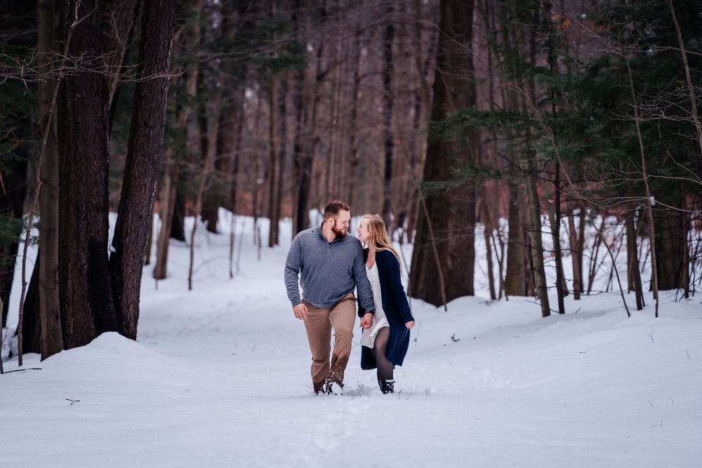 Winter engagement photo of couple walking through snowy forest path between tall pine trees.