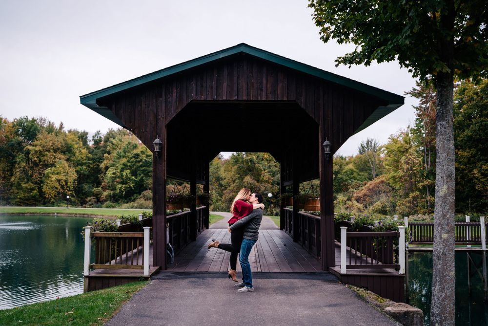 Couple sharing romantic moment on covered bridge over lake during fall engagement session.