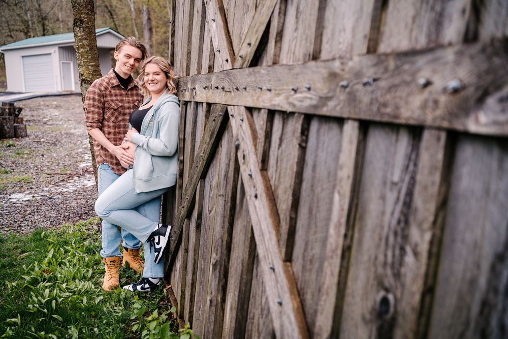 A couple stands beside a rustic wooden barn door wearing casual fall clothing and yellow boots.