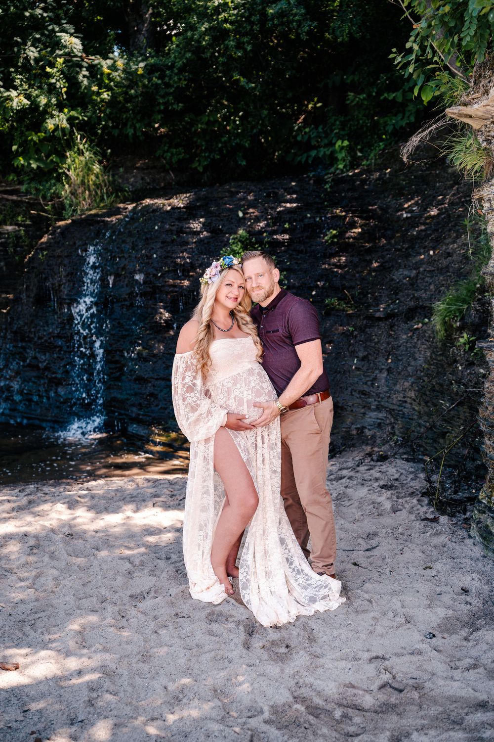 A maternity photoshoot featuring a couple posing by a small waterfall on a sandy beach.