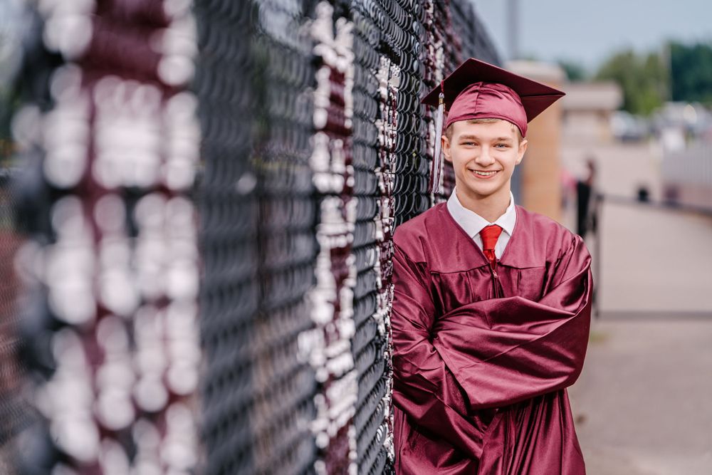 Graduate in maroon cap and gown poses against chain link fence with soft bokeh effect.