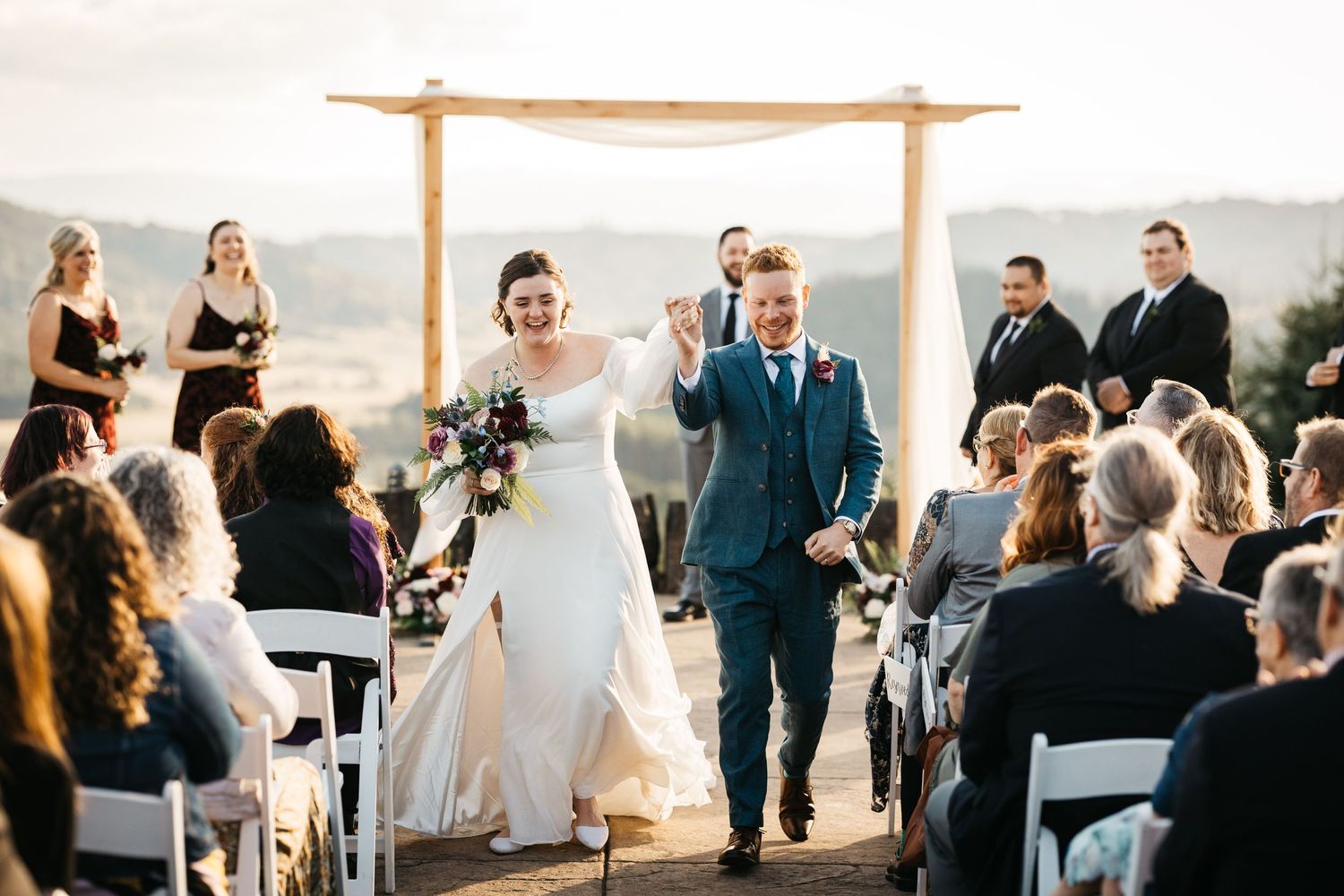 A joyful moment during a scenic outdoor wedding ceremony with guests seated and a wooden arch in the mountain backdrop.