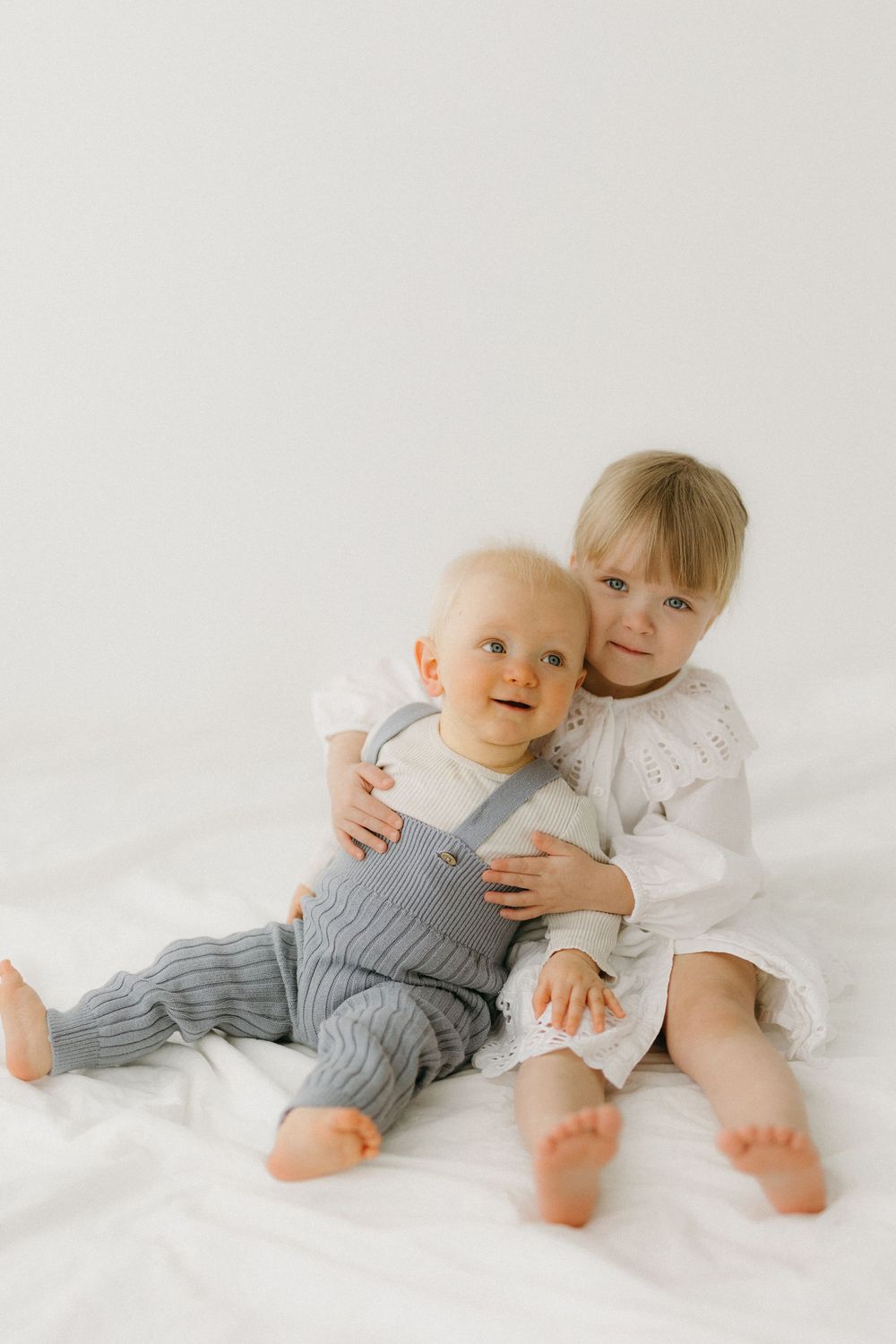 Two children in white and gray outfits lying on white bedding while playing together in a bright studio setting.
