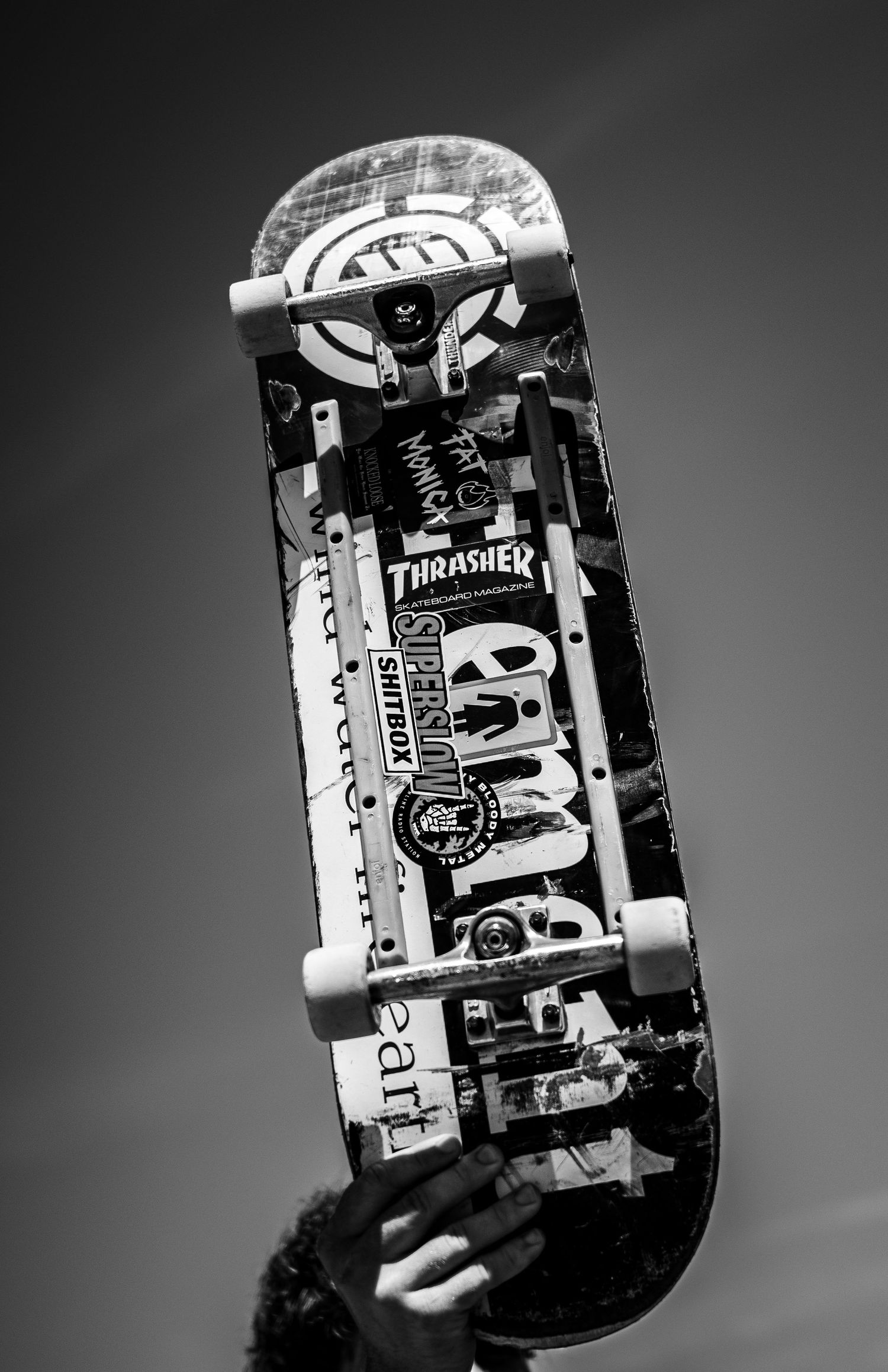 Black and white photo of a decorated skateboard held up against the sky, featuring Thrasher stickers.