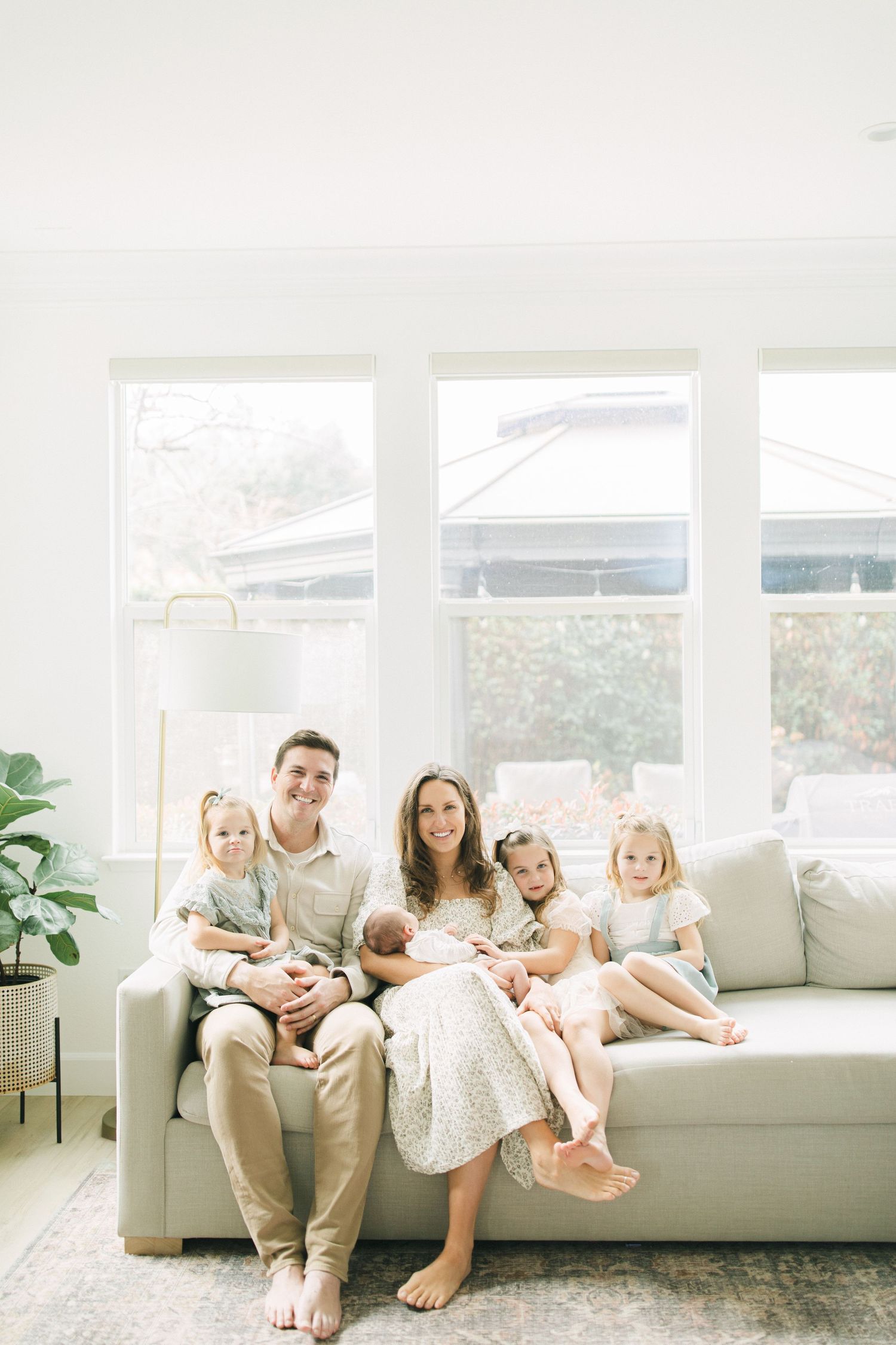 A family relaxes together on a light gray couch in a bright, airy room with white walls and large windows.
