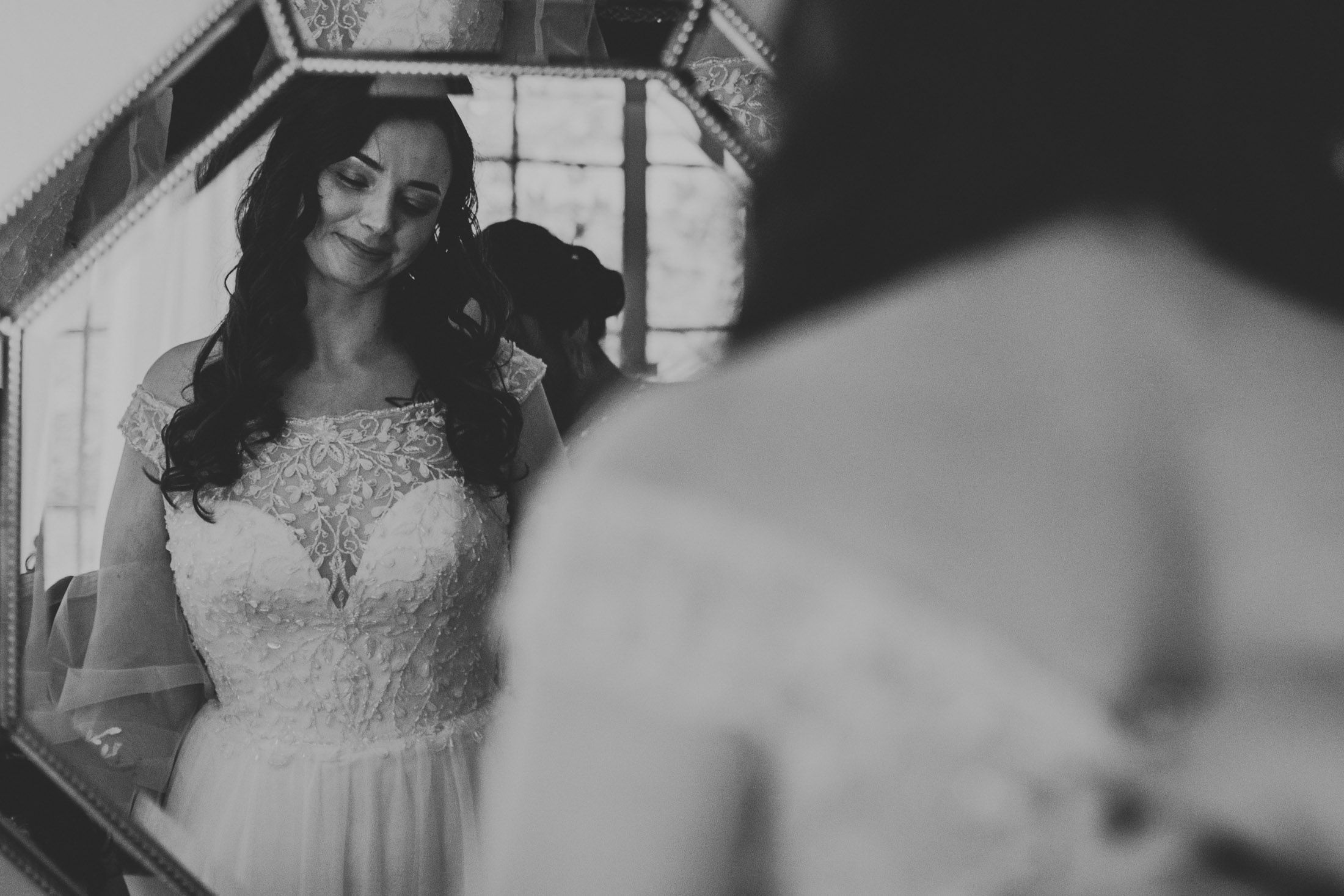 A black and white photograph shows someone in an elegant lace wedding dress standing near a window.