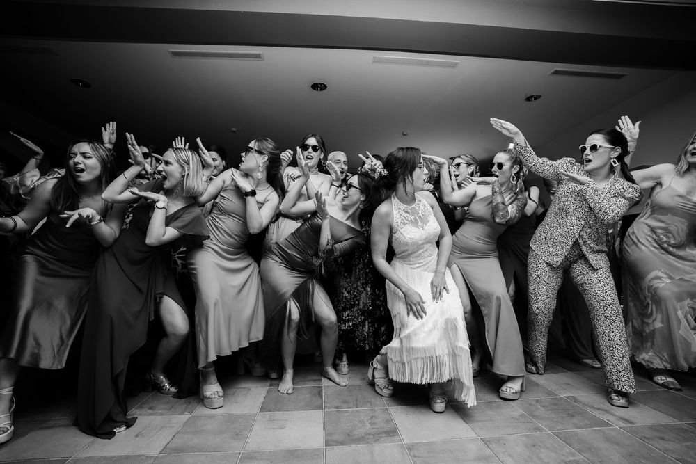 Black and white photo of wedding party guests dancing energetically in synchronized choreography at a reception.
