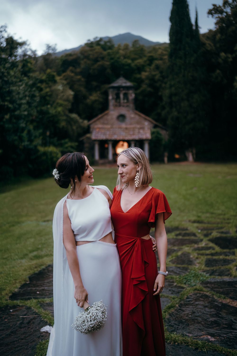 Two wedding guests in contrasting white and red dresses walk together on grassy grounds at sunset.