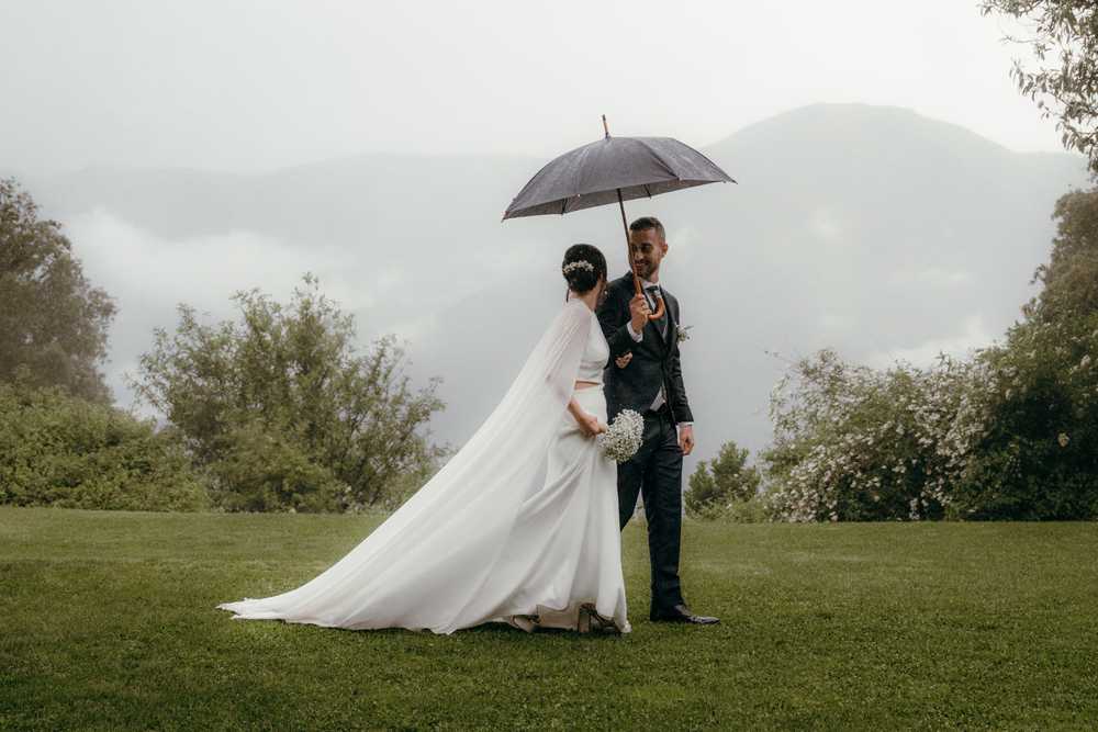 A couple with umbrellas share romantic wedding photos in the rain on a misty hillside.