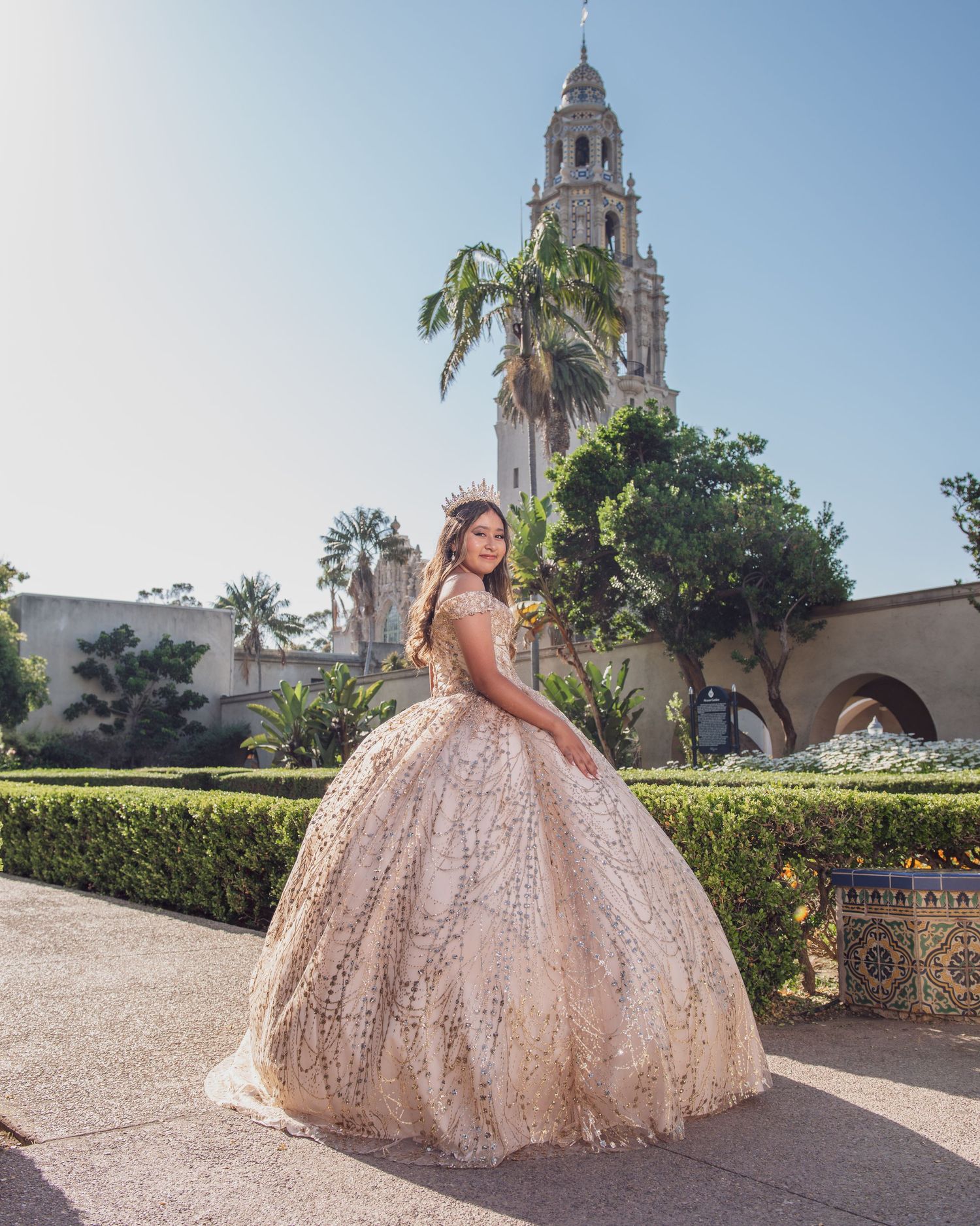 A stunning rose gold quinceañera ball gown sparkles in the sunlight against Spanish colonial architecture and palm trees.