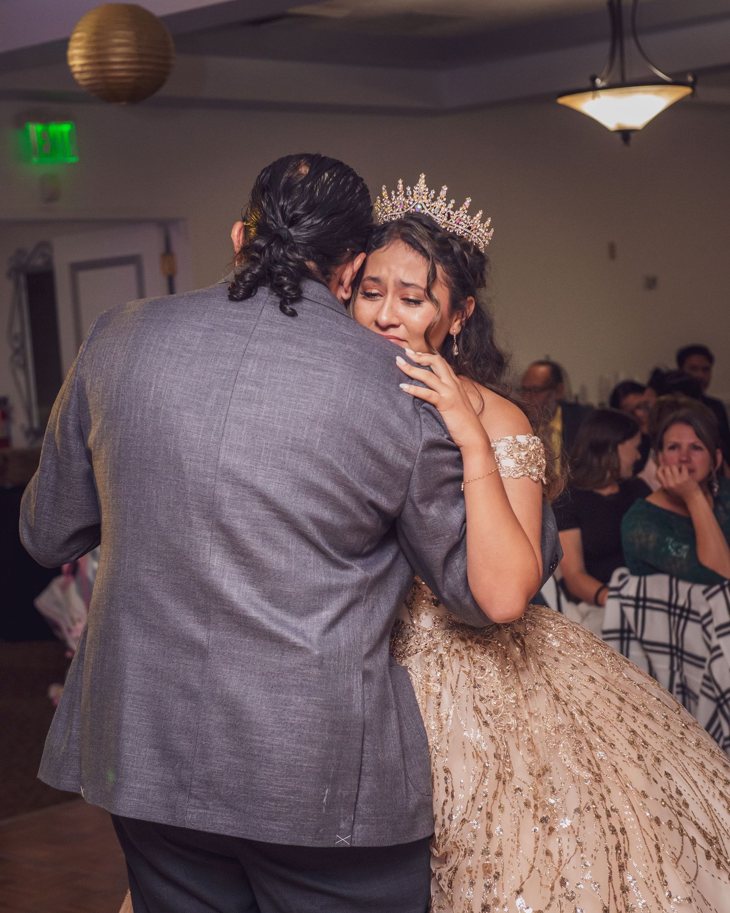 A quinceañera in a sparkly gown dances with someone wearing a grey suit at her celebration event.