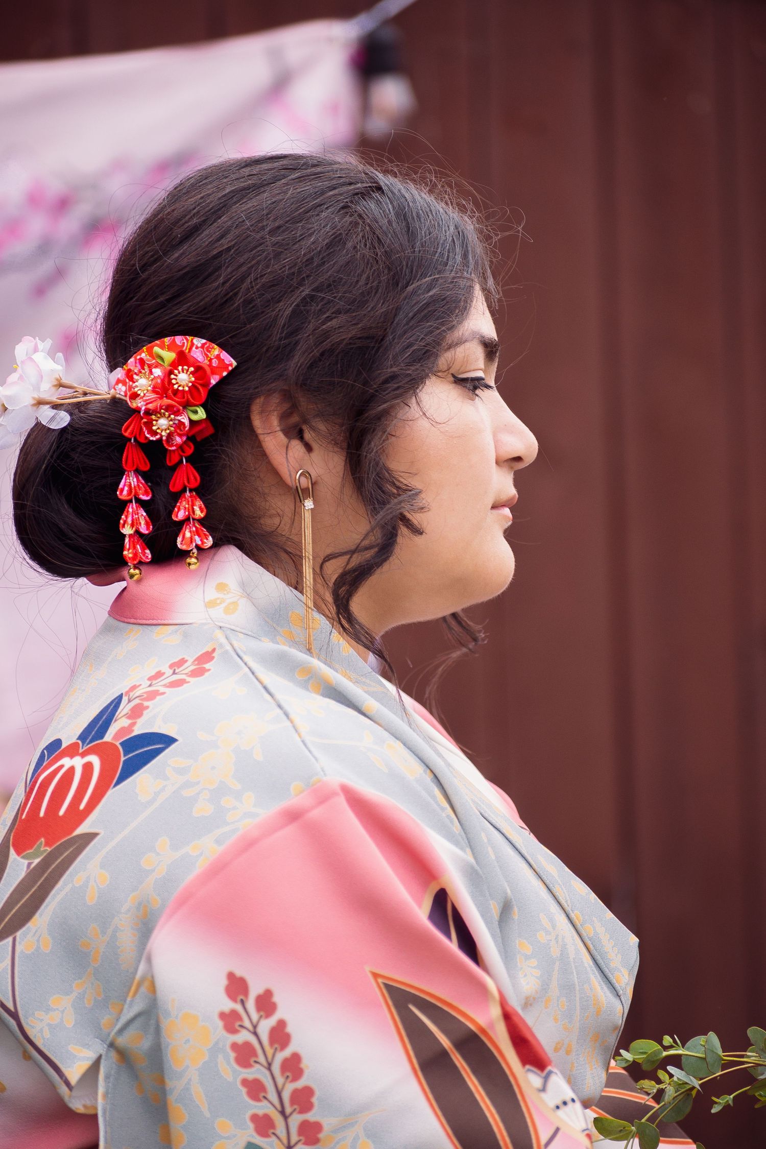 A woman in a traditional pink and blue floral kimono with red hair accessories poses in profile against a blurred background.