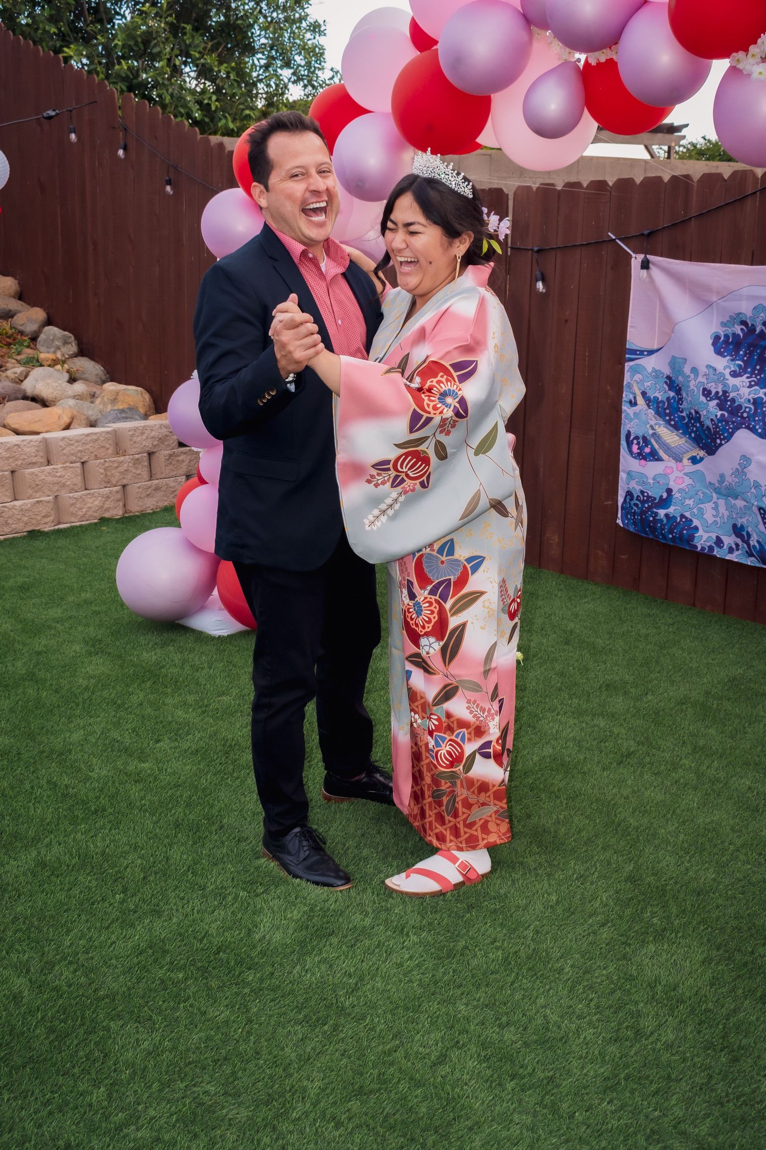 A couple shares a joyful dance moment on artificial grass under pink and red balloon arch decorations.