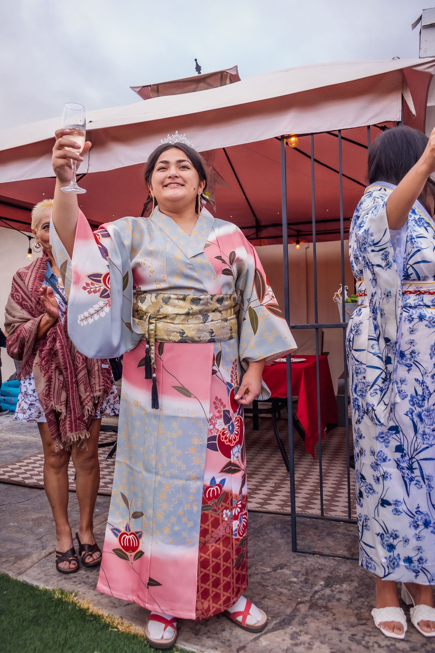 A person wearing a pink and gray patterned kimono raises a glass in celebration at an outdoor gathering.