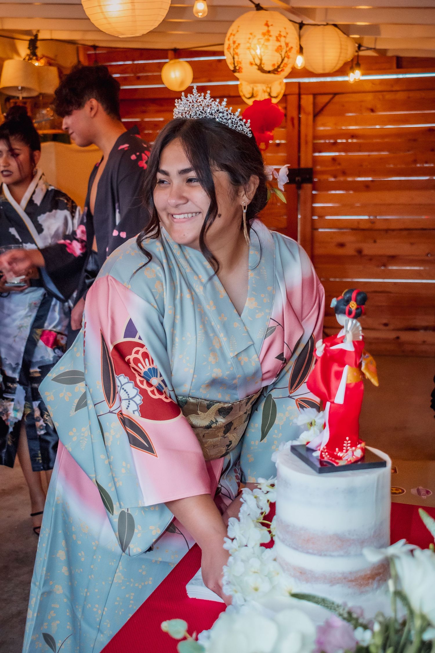 Couple in traditional dress cut their red and white wedding cake at a rustic venue with paper lantern lighting.