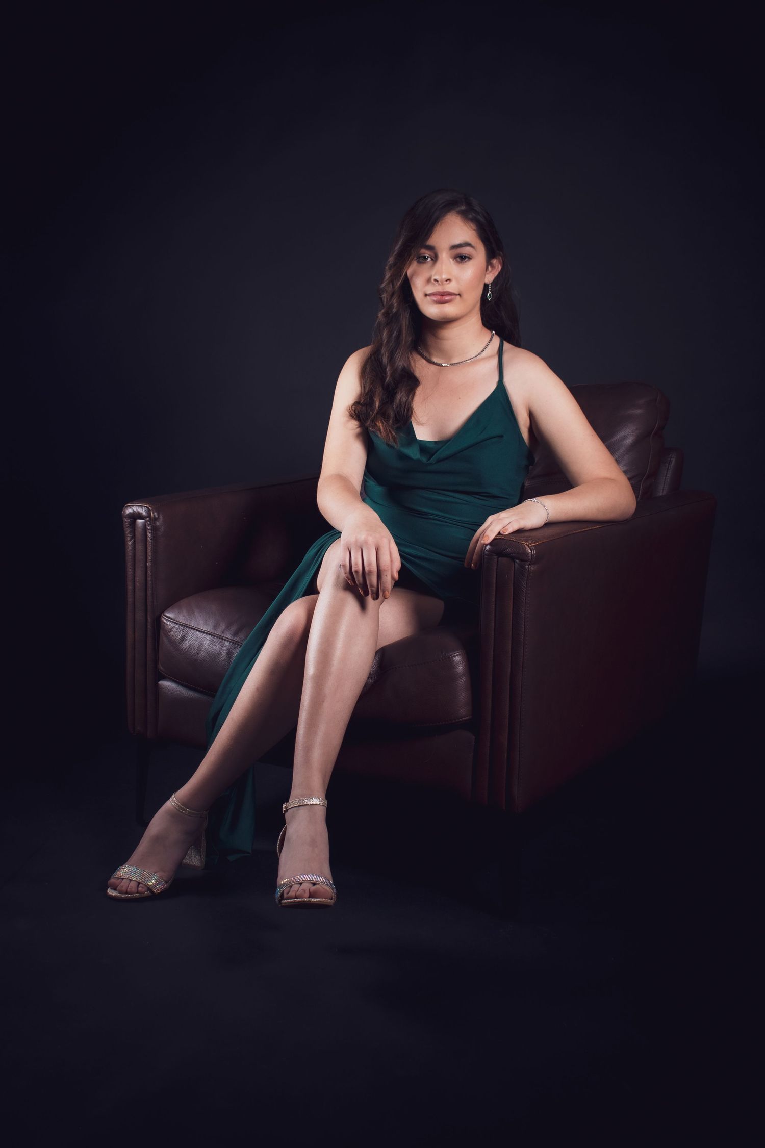 A person in a dark green dress poses elegantly on a leather chair against a black studio background.