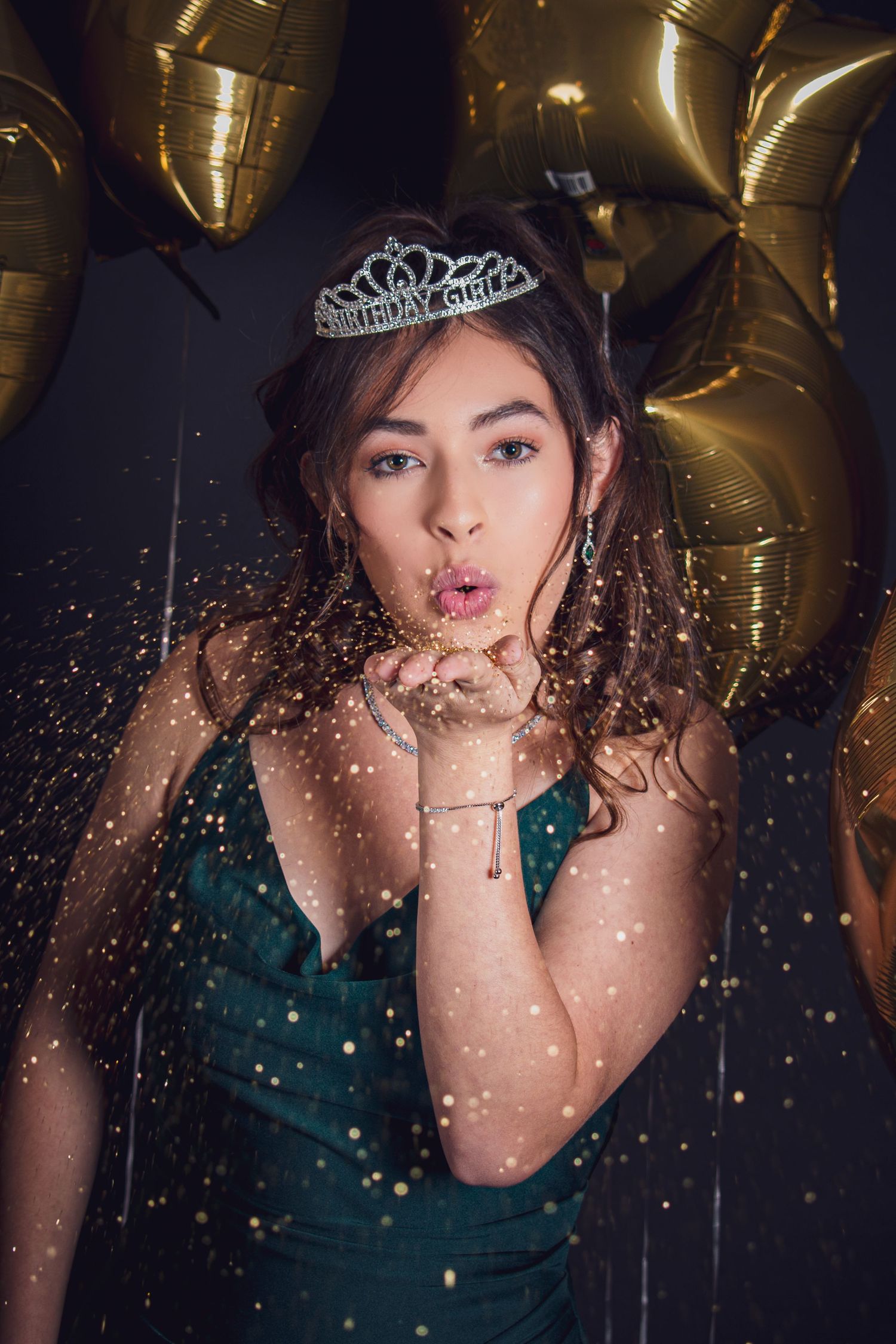 A person in a teal dress and tiara blows glitter while posing against a dark background with gold balloons.