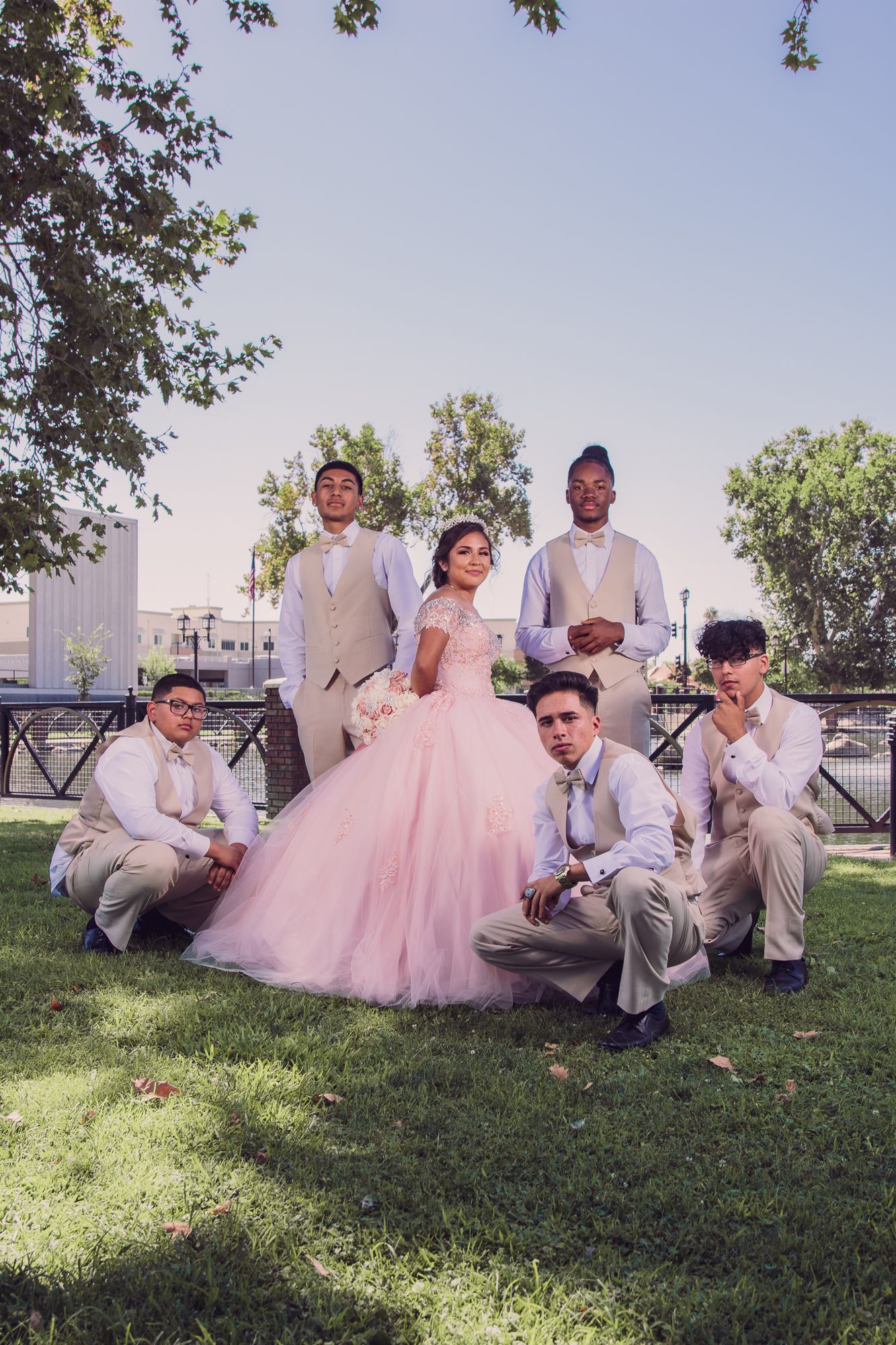 A group in formal attire poses on grass, with a person wearing a pink ball gown as the centerpiece.