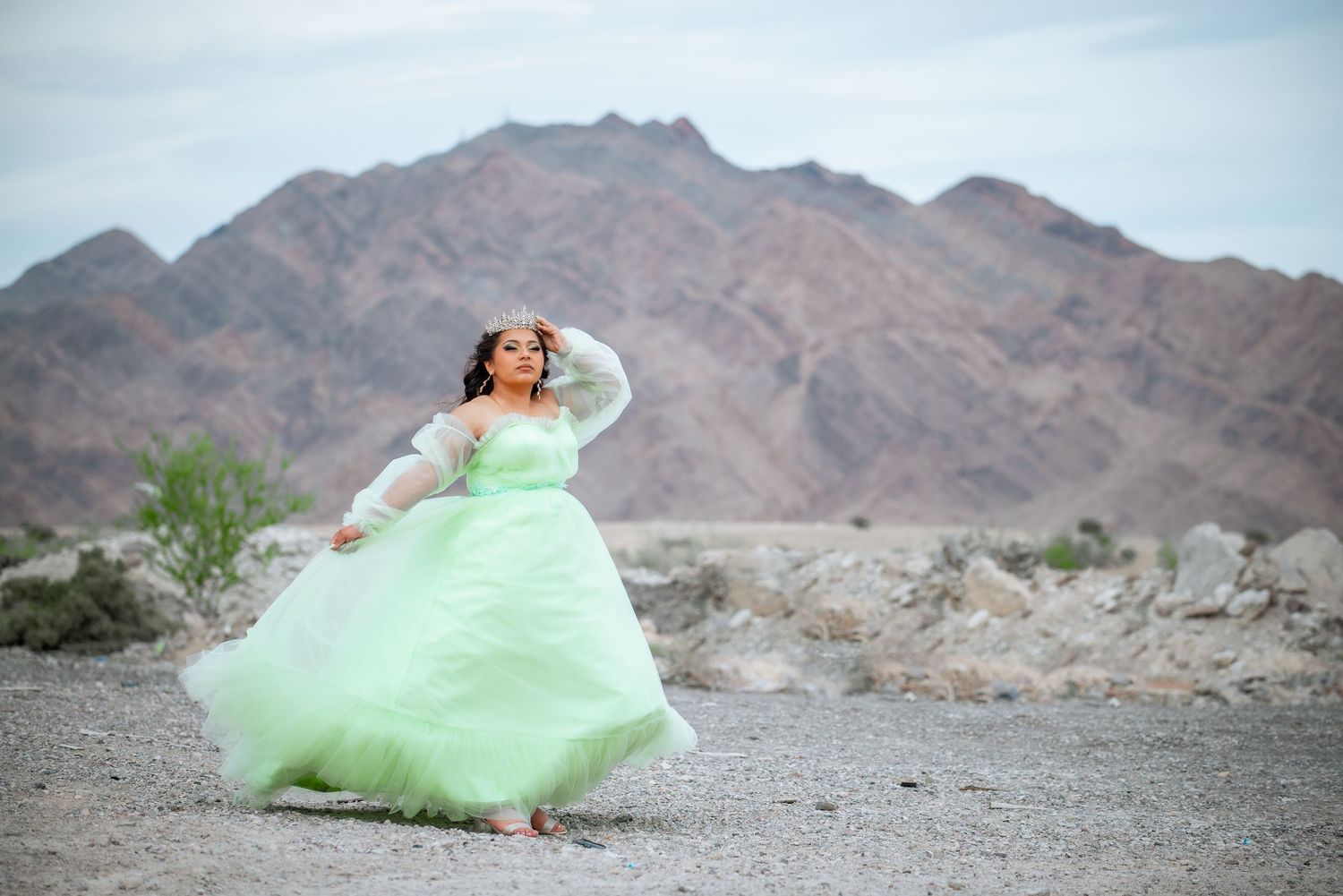A person in a mint green gown poses dramatically against a desert mountain landscape at sunset.