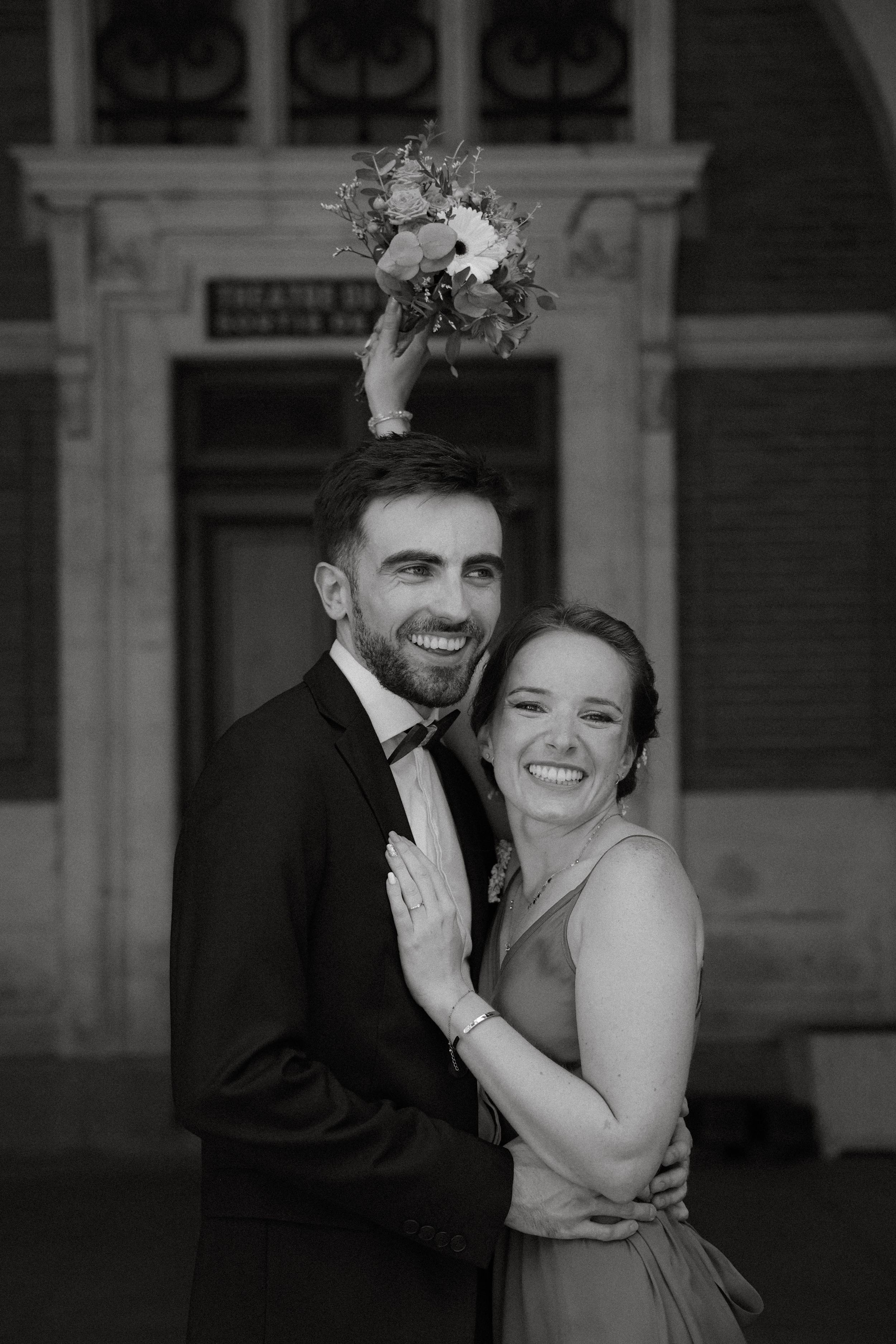 Black and white portrait of smiling newlyweds embracing with bouquet tossed above them.