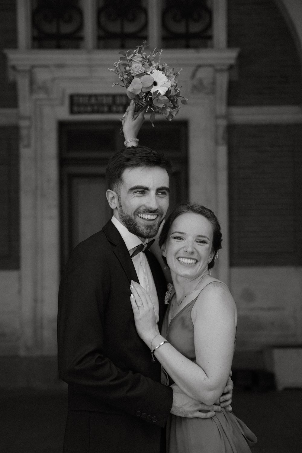Black and white portrait of smiling newlyweds embracing with bouquet tossed above them.