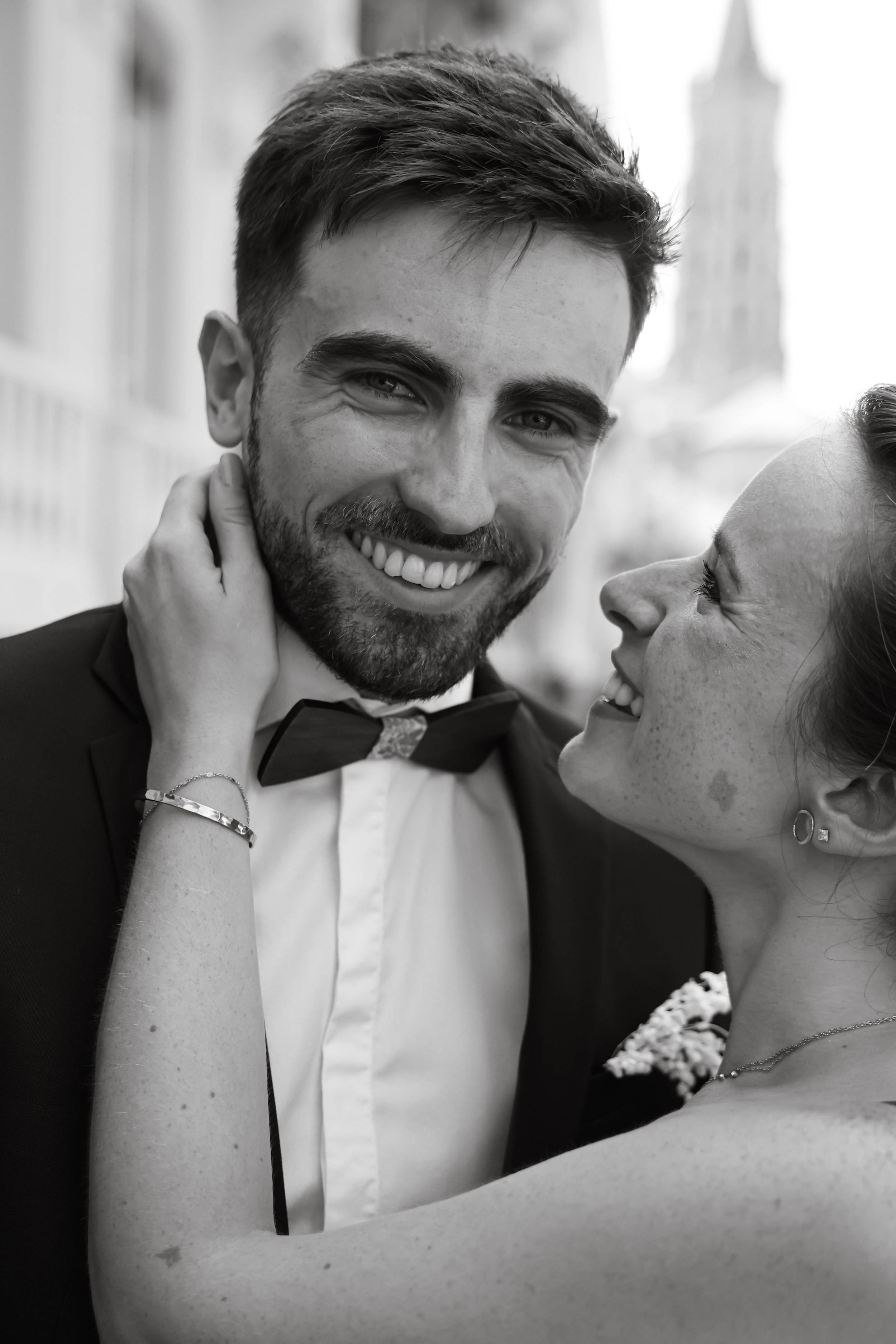 Black and white portrait of smiling groom in bow tie with bride embracing him tenderly.