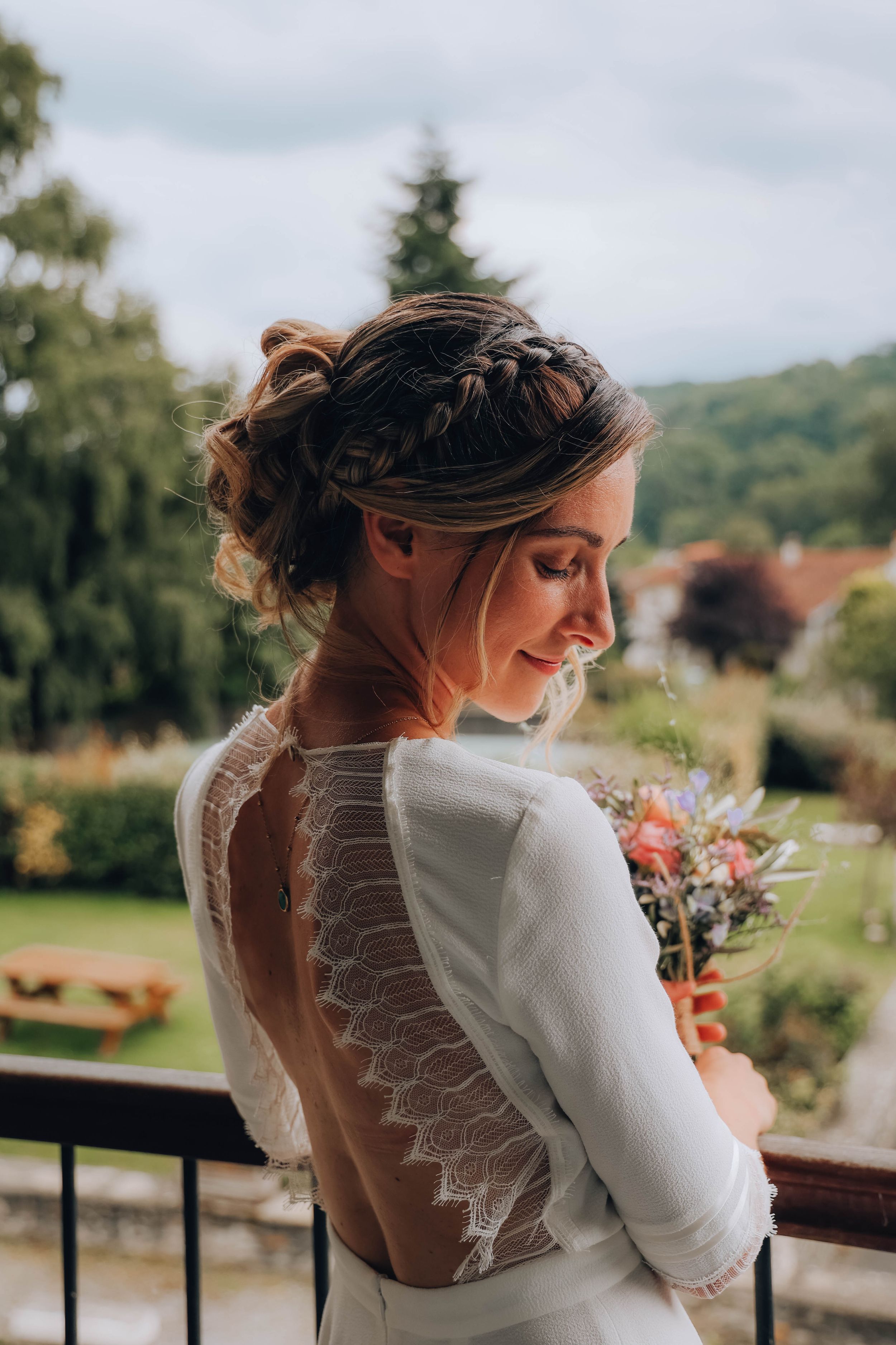 Bride in backless lace wedding dress with braided updo holds bouquet on balcony with garden view.