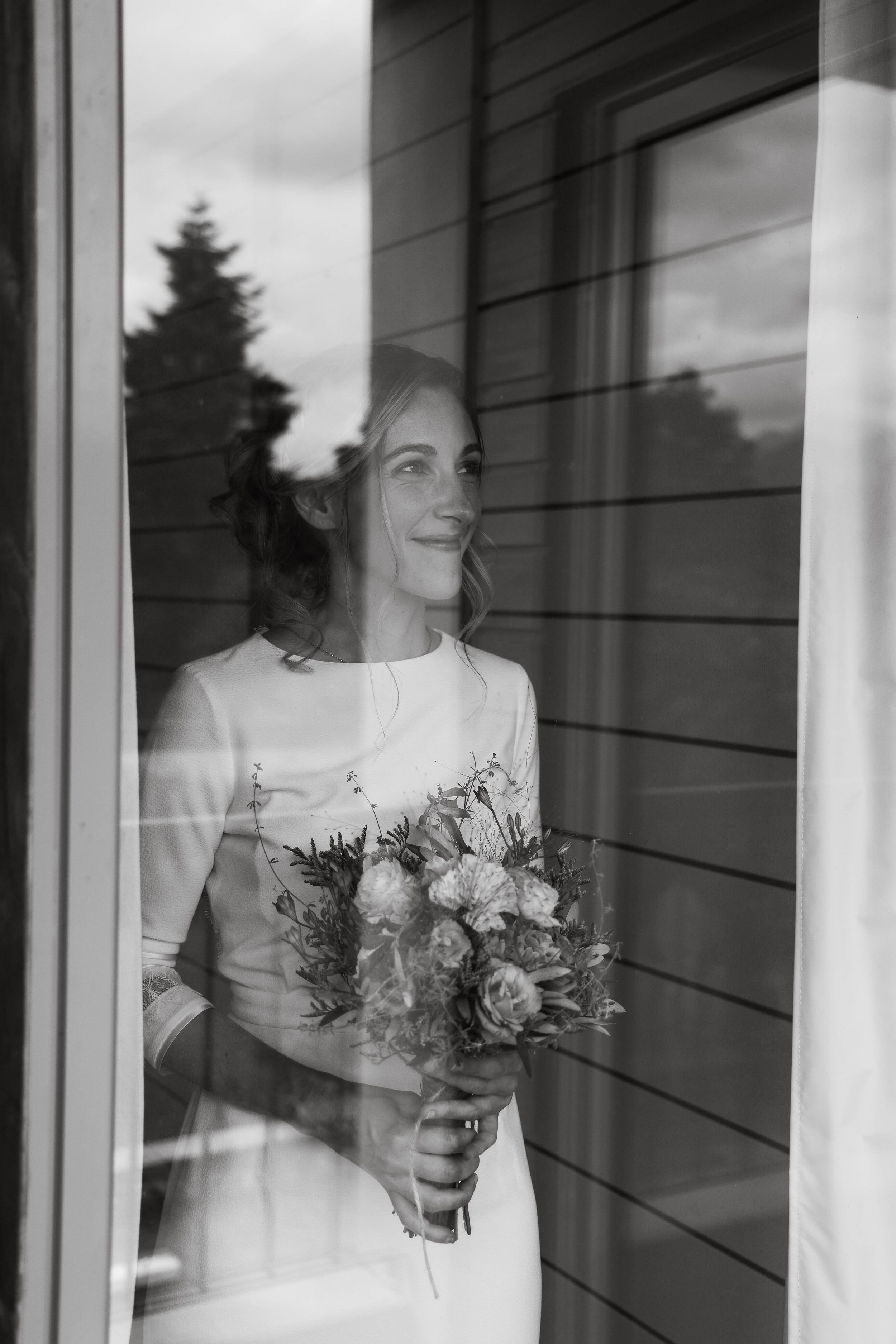 Black and white photo of a smiling bride holding a bouquet, gazing out a window in her wedding dress.