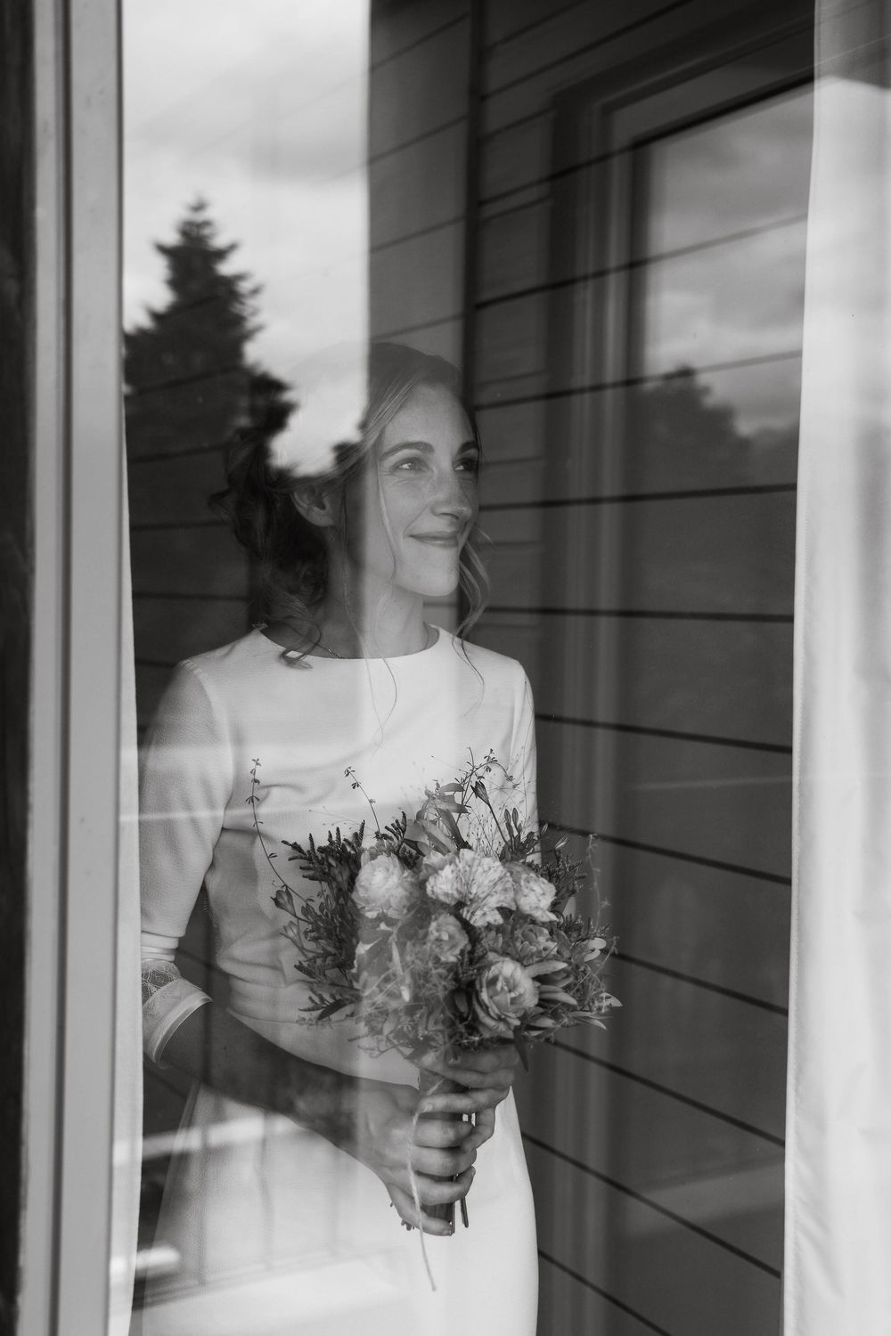Black and white photo of a smiling bride holding a bouquet, gazing out a window in her wedding dress.