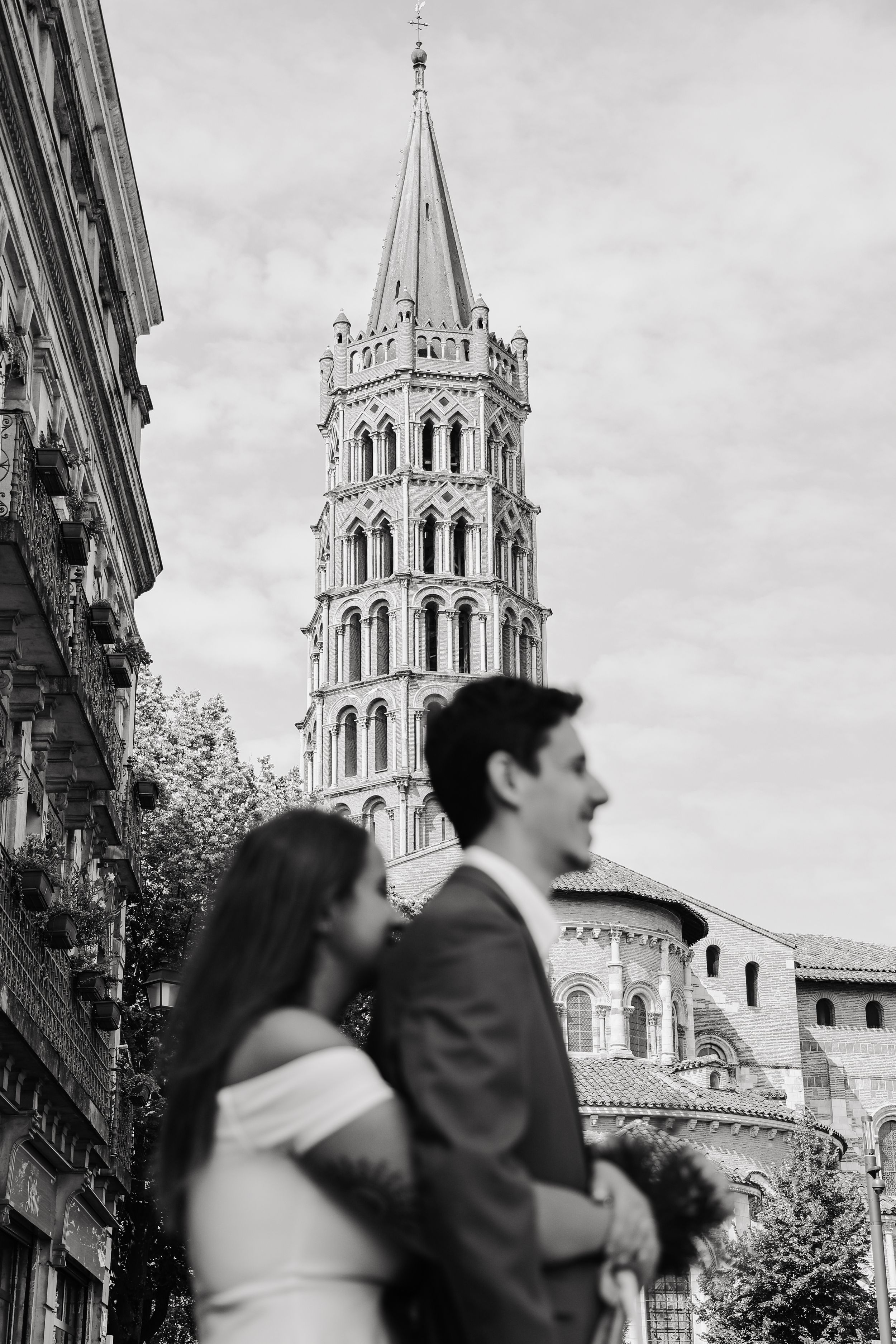 Black and white photo of wedding couple standing before iconic Toulouse church bell tower.
