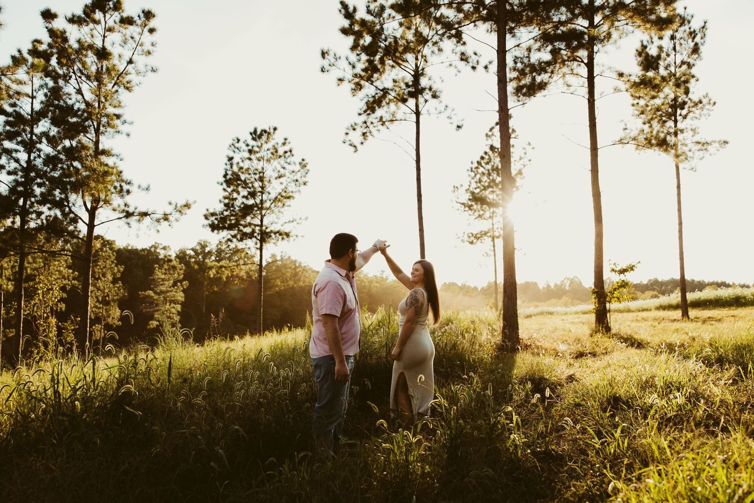 Couple dancing in golden sunlit meadow surrounded by tall pine trees during romantic outdoor engagement session.