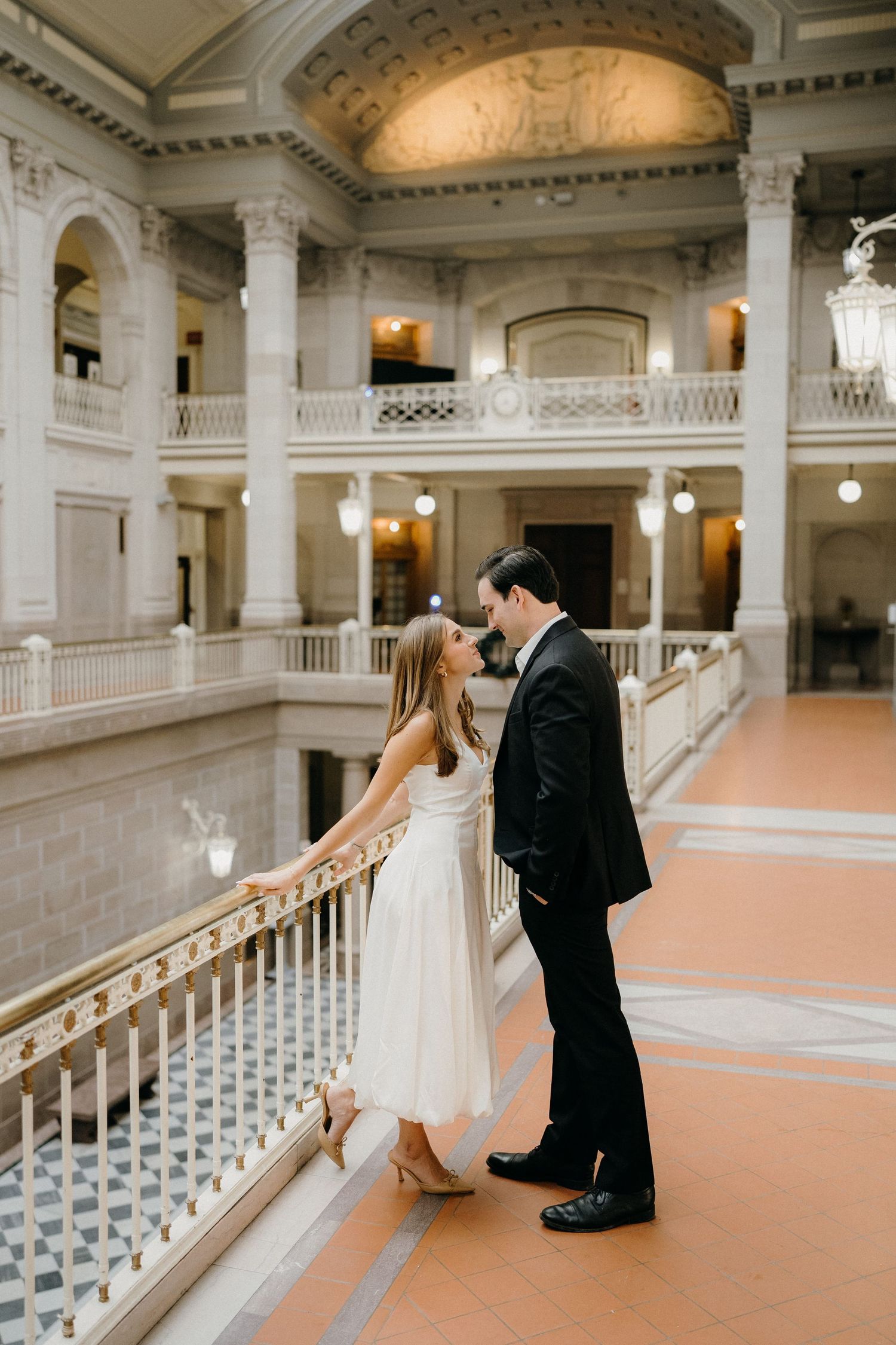 A bride and groom share romantic moments in a grand marble hall with elegant columns and balustrades.