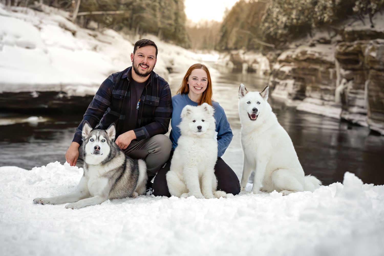 A man and woman sitting in the snow with two white dogs.