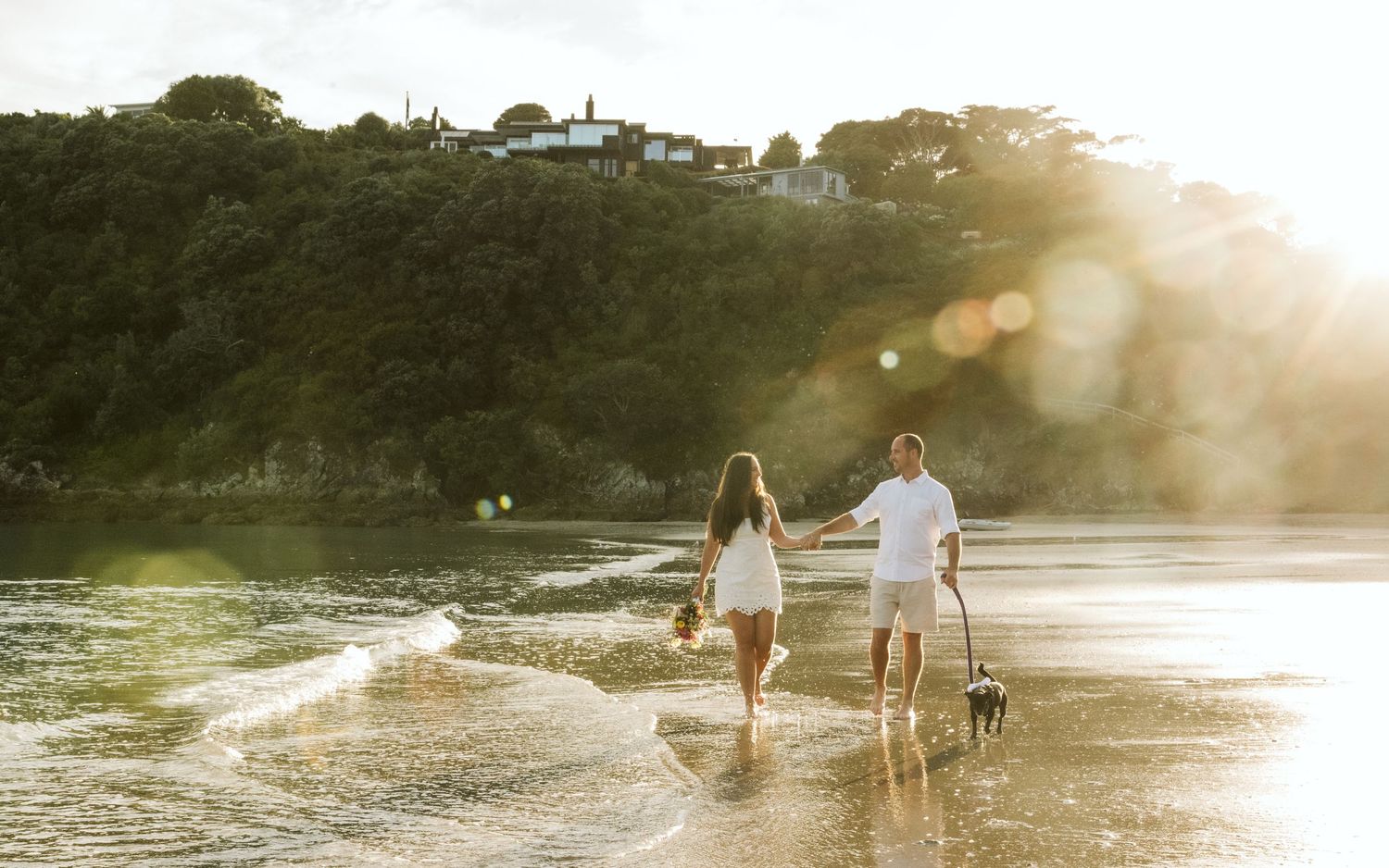 A couple in white and pink attire poses joyfully in a green outdoor setting on a cloudy day.