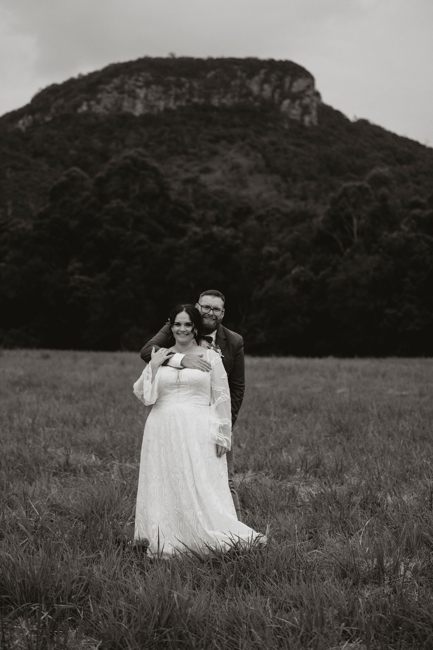 A couple in wedding attire walking through a field while birds fly overhead in black and white.
