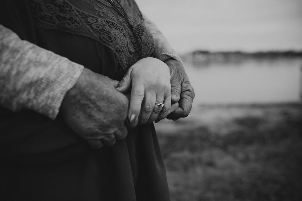 A close-up black and white photo shows two hands gently clasped together against a blurred outdoor background.