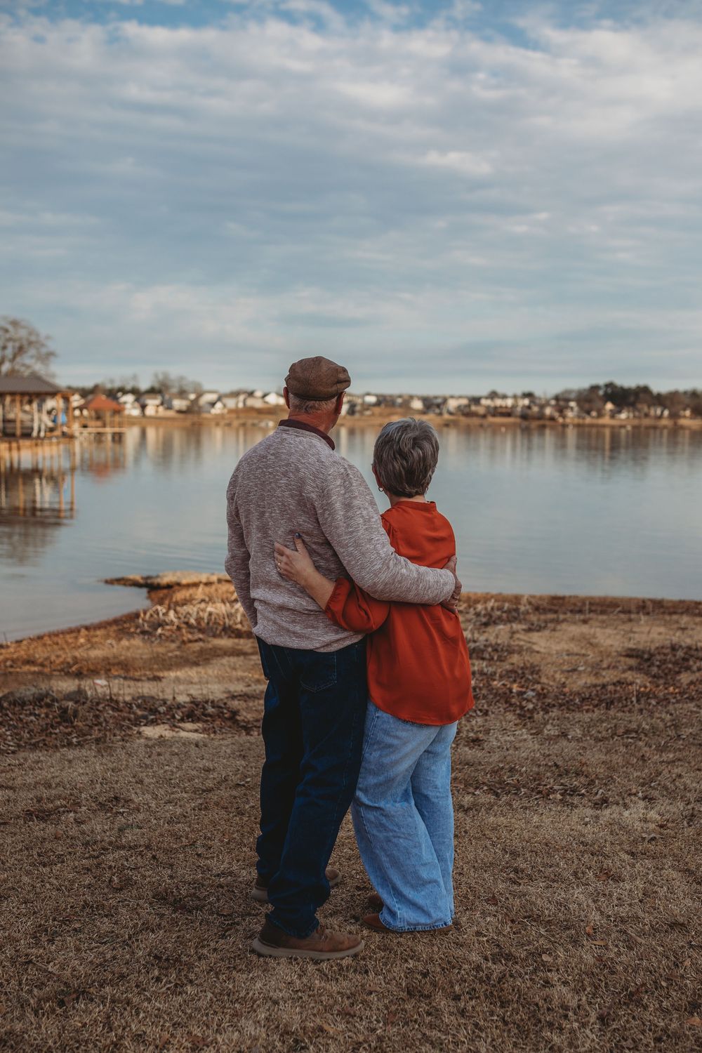 Two people sit close together on the shoreline overlooking a calm lake with houses visible in the distance.
