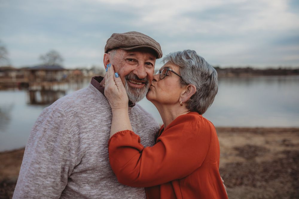 Elderly couple embracing on a beach at sunset with a pier in the background.