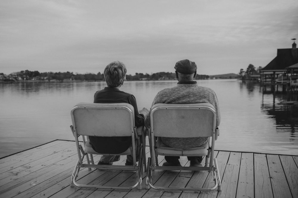 Two people sit in lawn chairs on a wooden dock overlooking a serene lake in black and white.