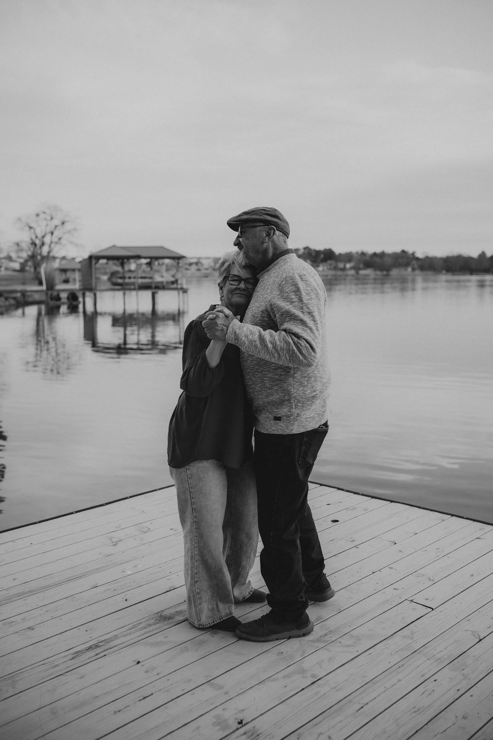 A couple embraces on a wooden dock overlooking a serene lake in a series of black and white photographs.