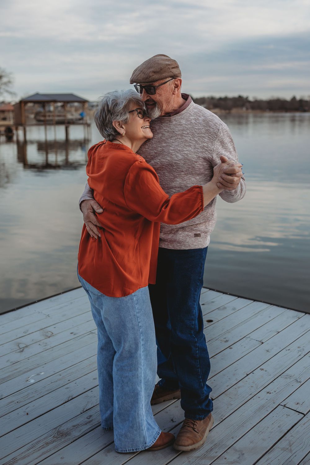 A couple in casual attire embraces on a wooden dock overlooking a serene lake during sunset.