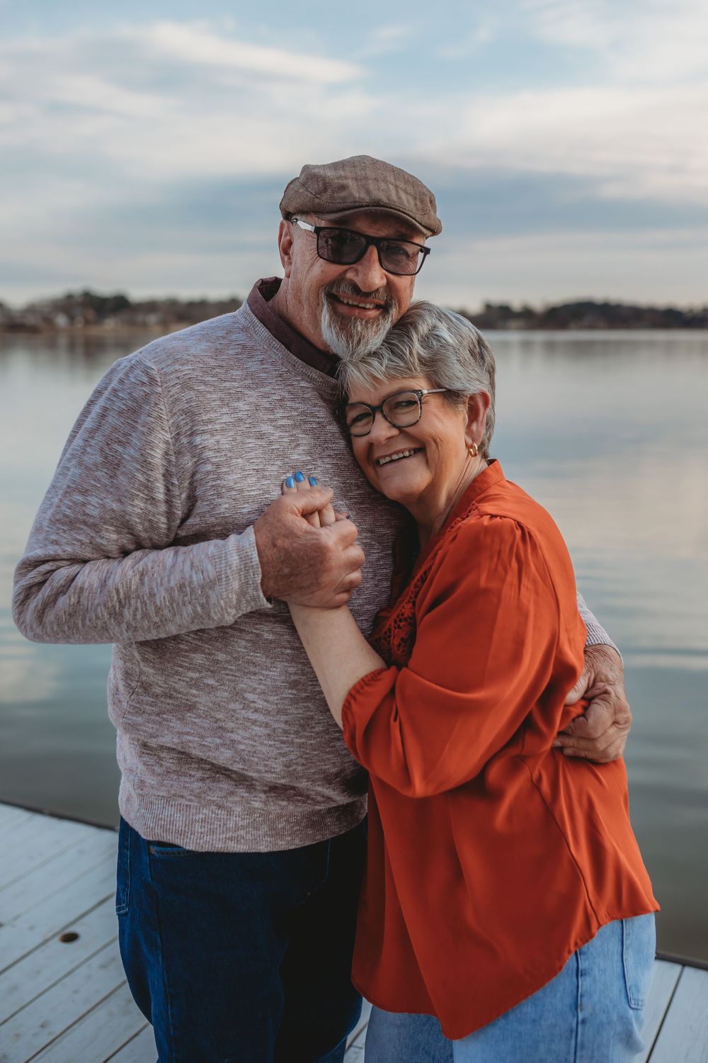 A couple embraces and laughs together on a wooden dock by a serene lake on a cloudy day.