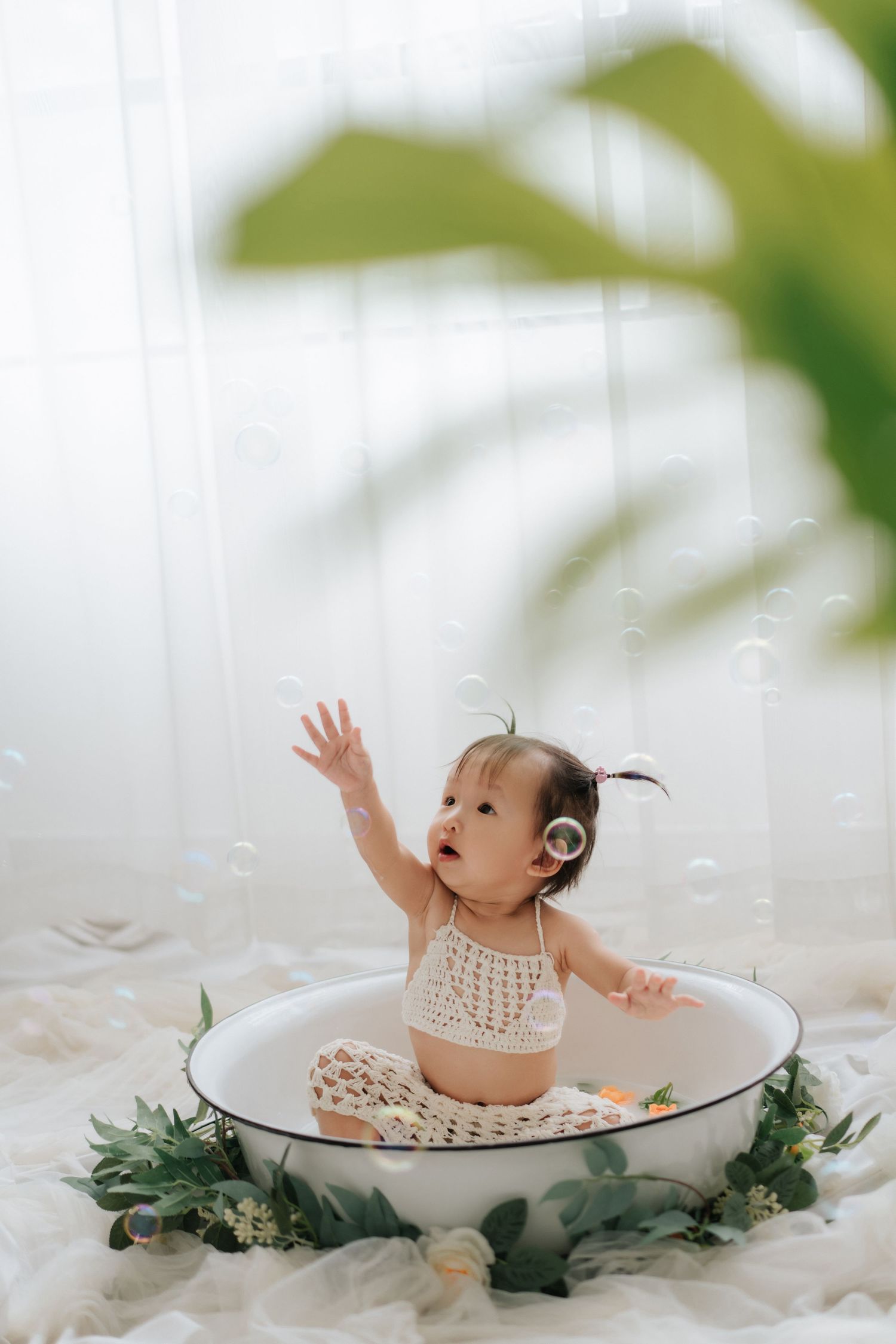 Baby sits on wooden floor surrounded by teal and white balloons during first birthday photoshoot.