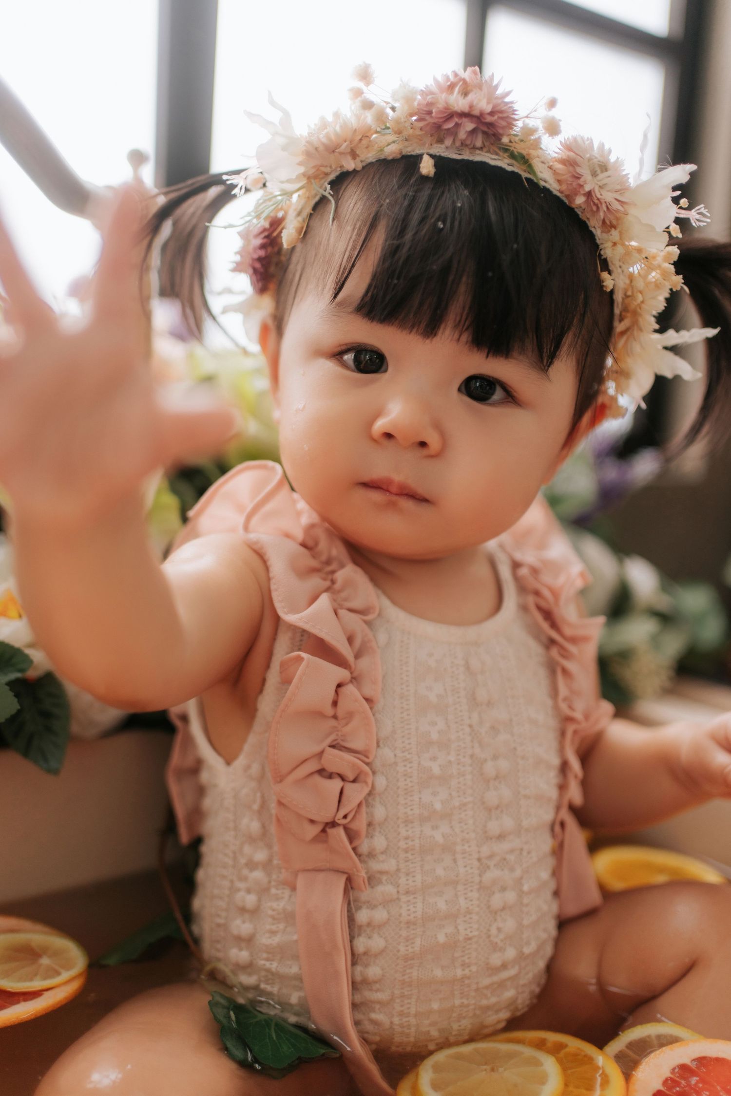 Baby wearing pink knit sweater and floral headband sits near citrus fruits in warm natural lighting.