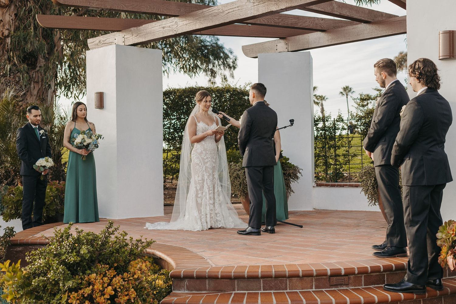 A couple exchanges vows during an outdoor wedding ceremony surrounded by green and white columns at San Clemente Shores.