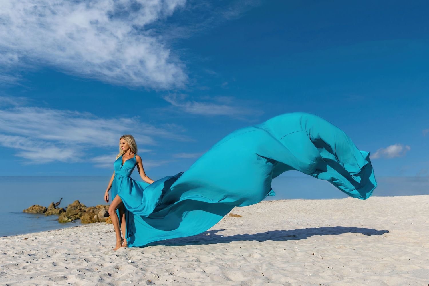 A flowing turquoise fabric creates a dynamic wave-like motion against a bright blue sky and white sandy beach background.