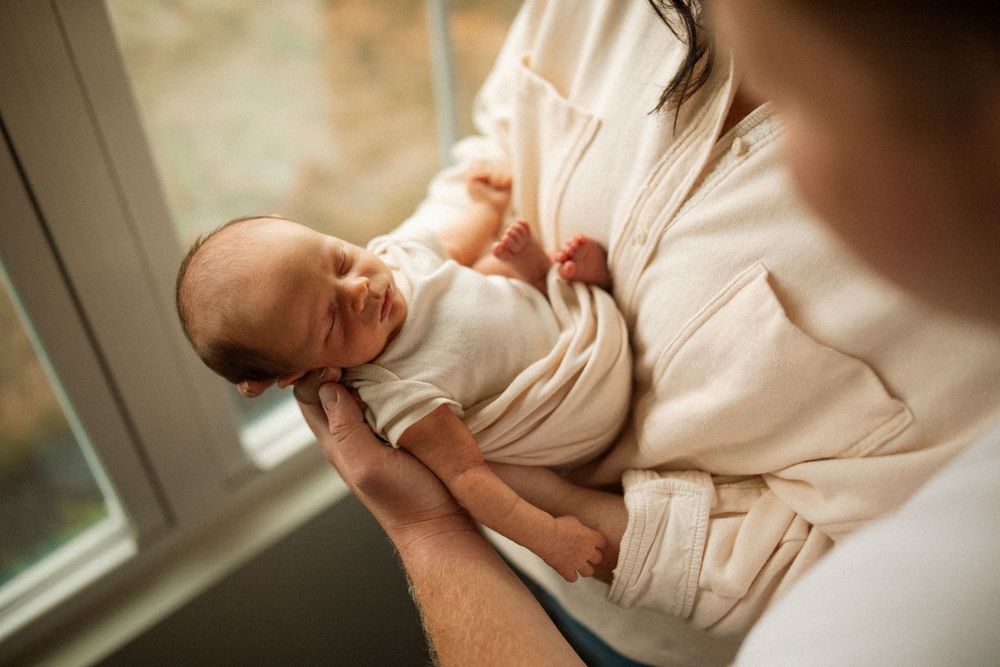 A newborn baby wrapped in soft white fabric is cradled in someone's arms near a window.