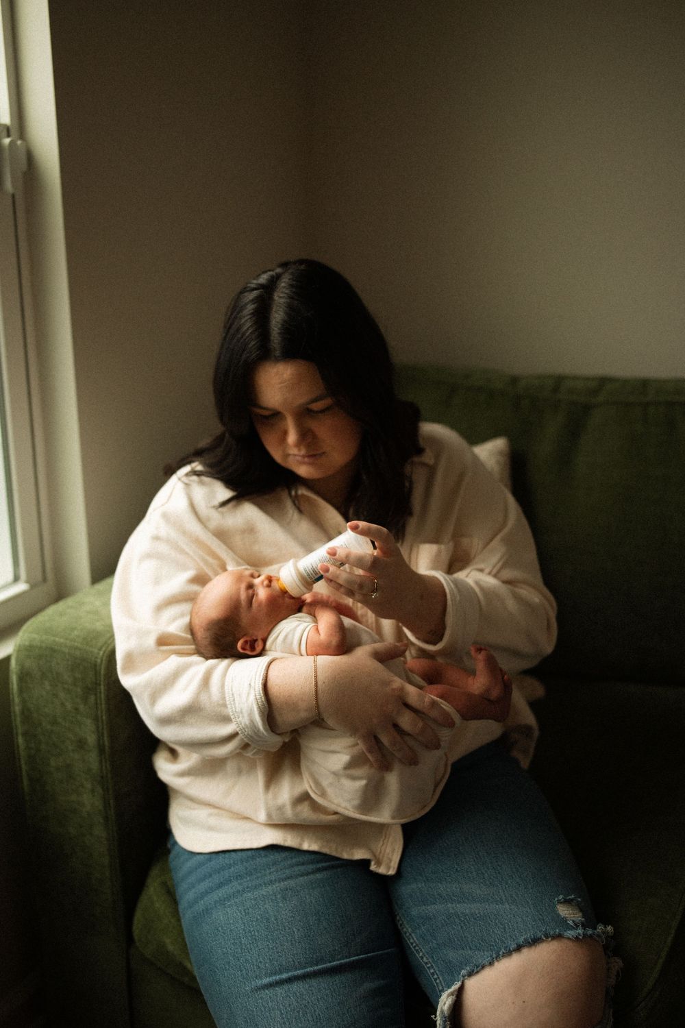 A parent in a white sweater tenderly feeds a baby while sitting on a green couch near a window.