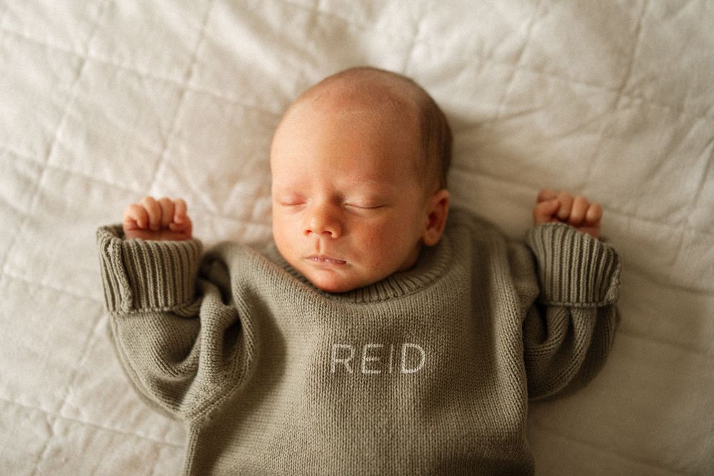 A sleeping newborn wearing a cozy gray knit sweater lies on white bedding with arms outstretched.