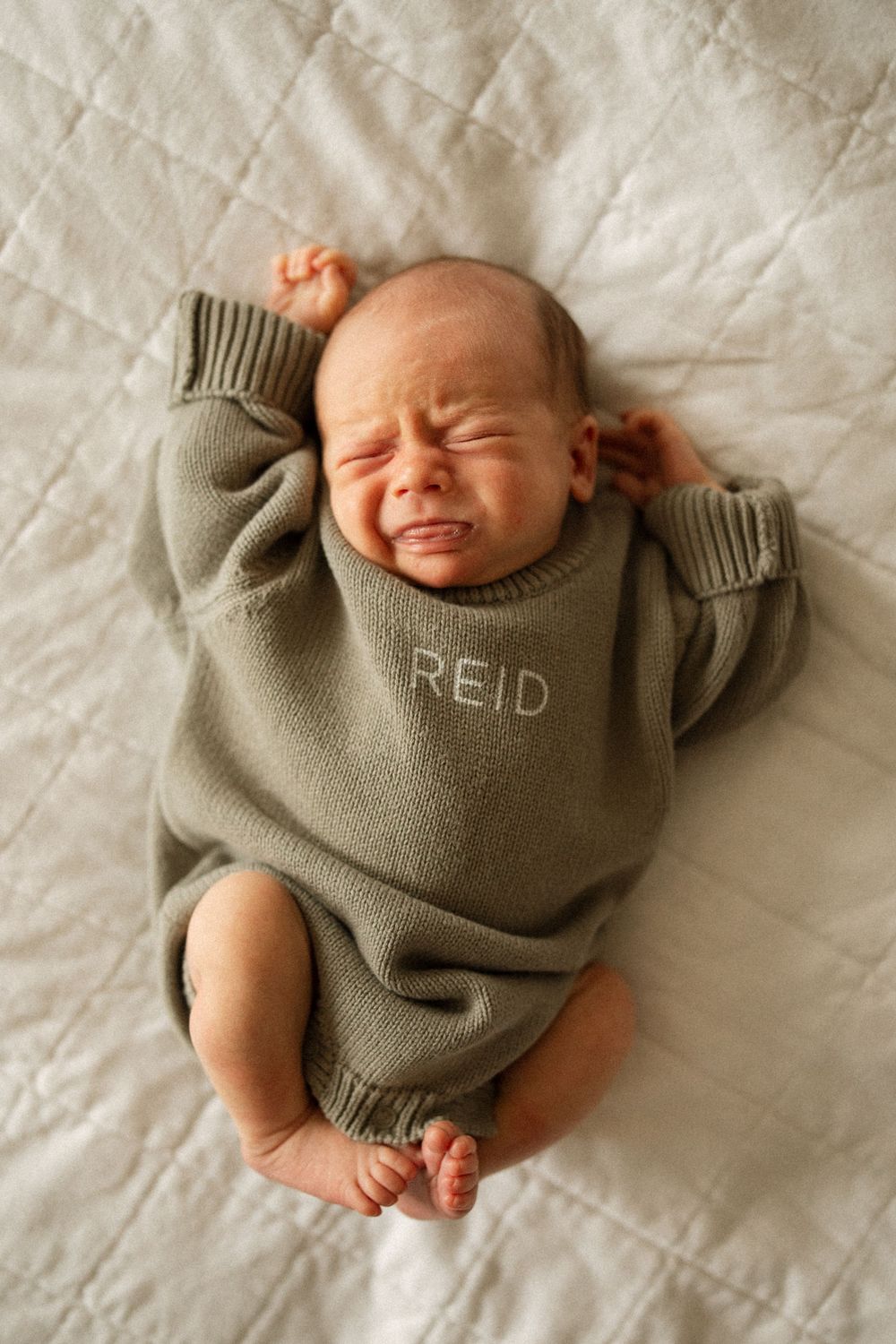 A newborn baby wearing a cozy gray sweater with 'Reid' embroidered lies on white bedding in multiple sweet poses.